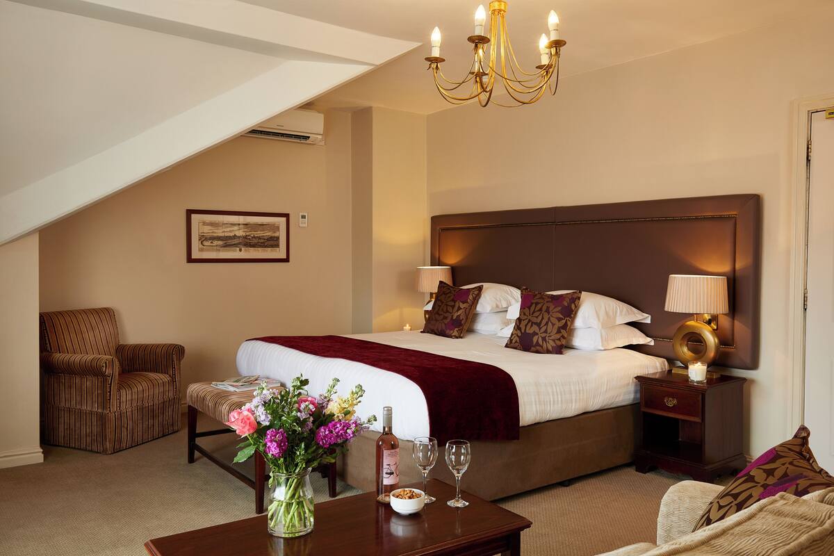 A hotel room with a large bed, a chair, a table with a vase of flowers, and a chandelier.