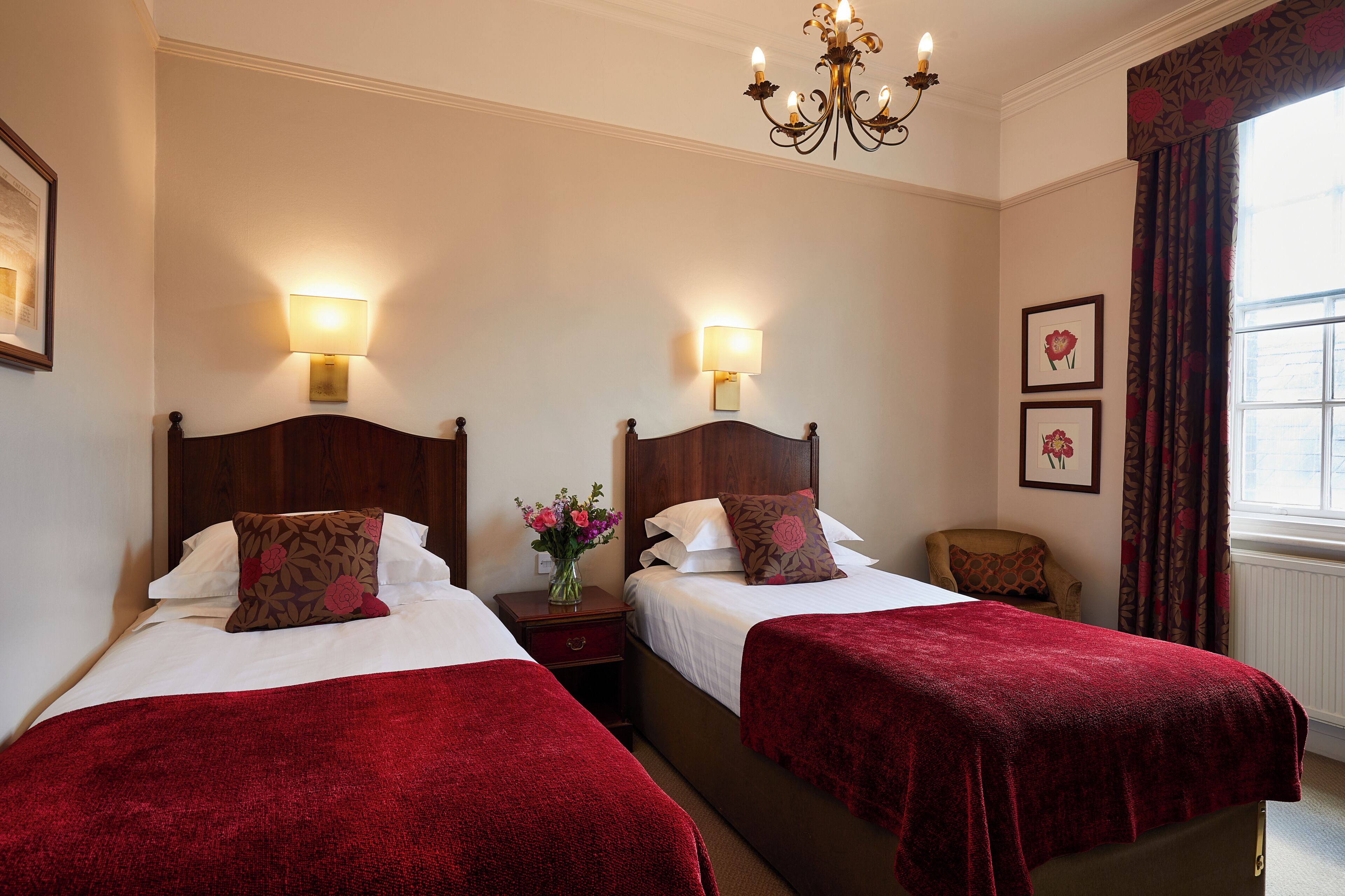 A hotel room with two beds, a nightstand, a vase with flowers, a chair, and a window with curtains.