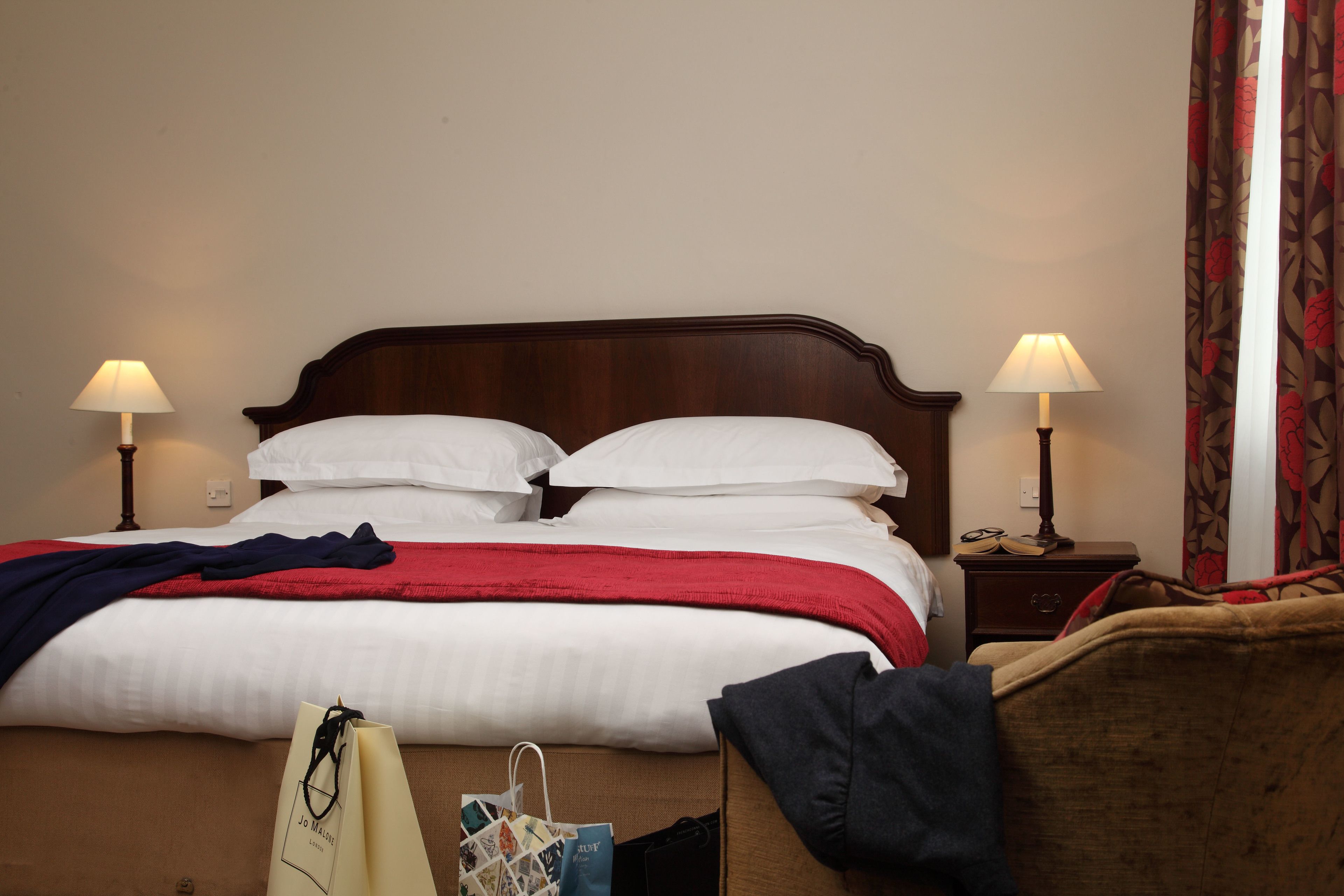A neatly made bed with white linens and a red blanket, two bedside lamps, a wooden nightstand with a lamp, and a brown armchair.
