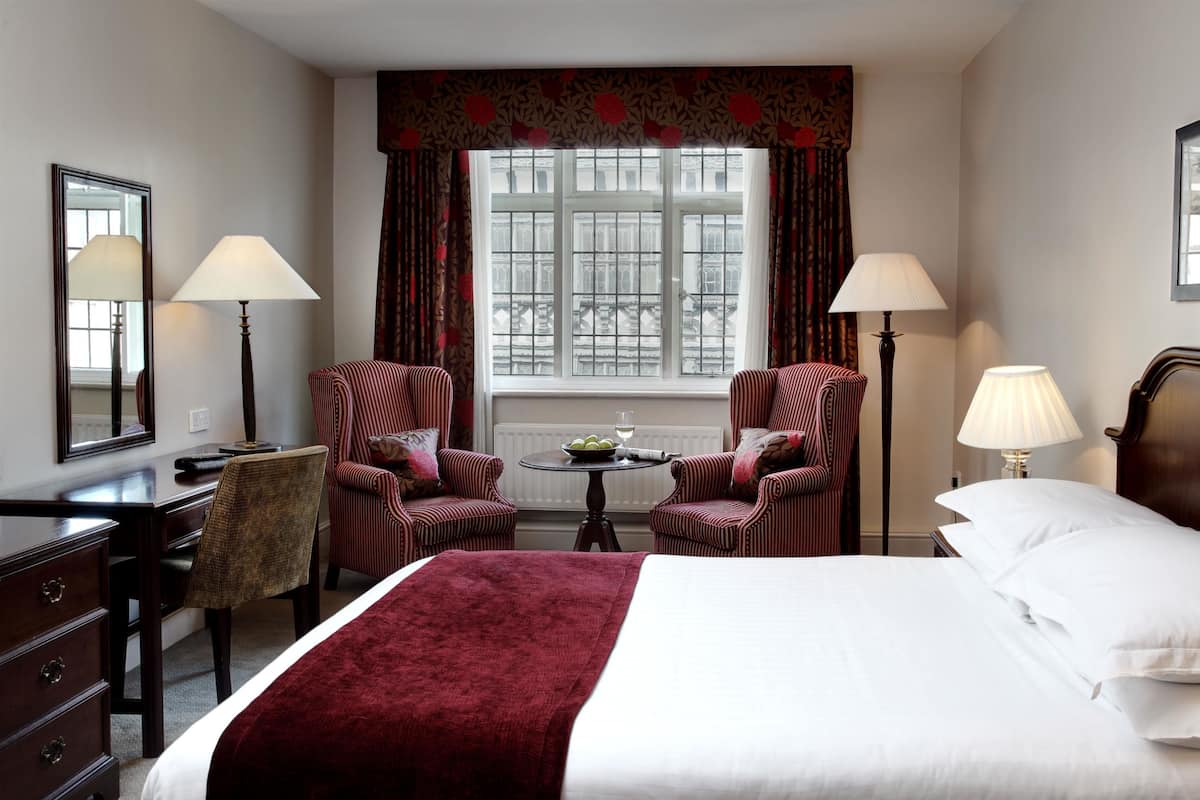 A hotel room with a bed, two armchairs, a small table with a bowl of fruit and a glass of wine, a desk with a lamp, and a window with curtains.