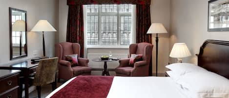 Una habitación de hotel con una cama, dos sillones, una mesita con un frutero y una copa de vino, un escritorio con una lámpara, y una ventana con cortinas.