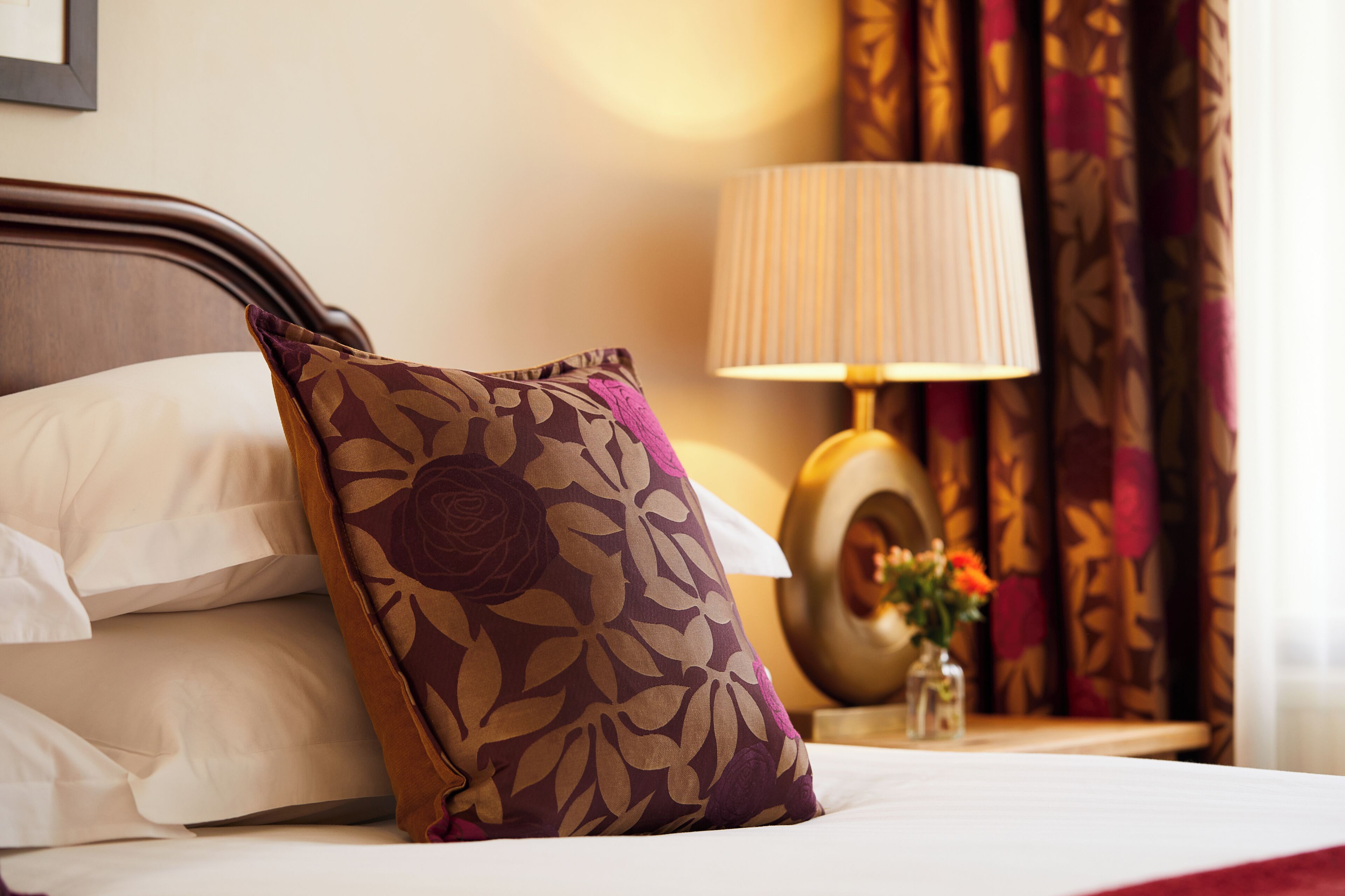 A neatly made bed with a floral pillow, a bedside lamp, and a vase with flowers on a nightstand.