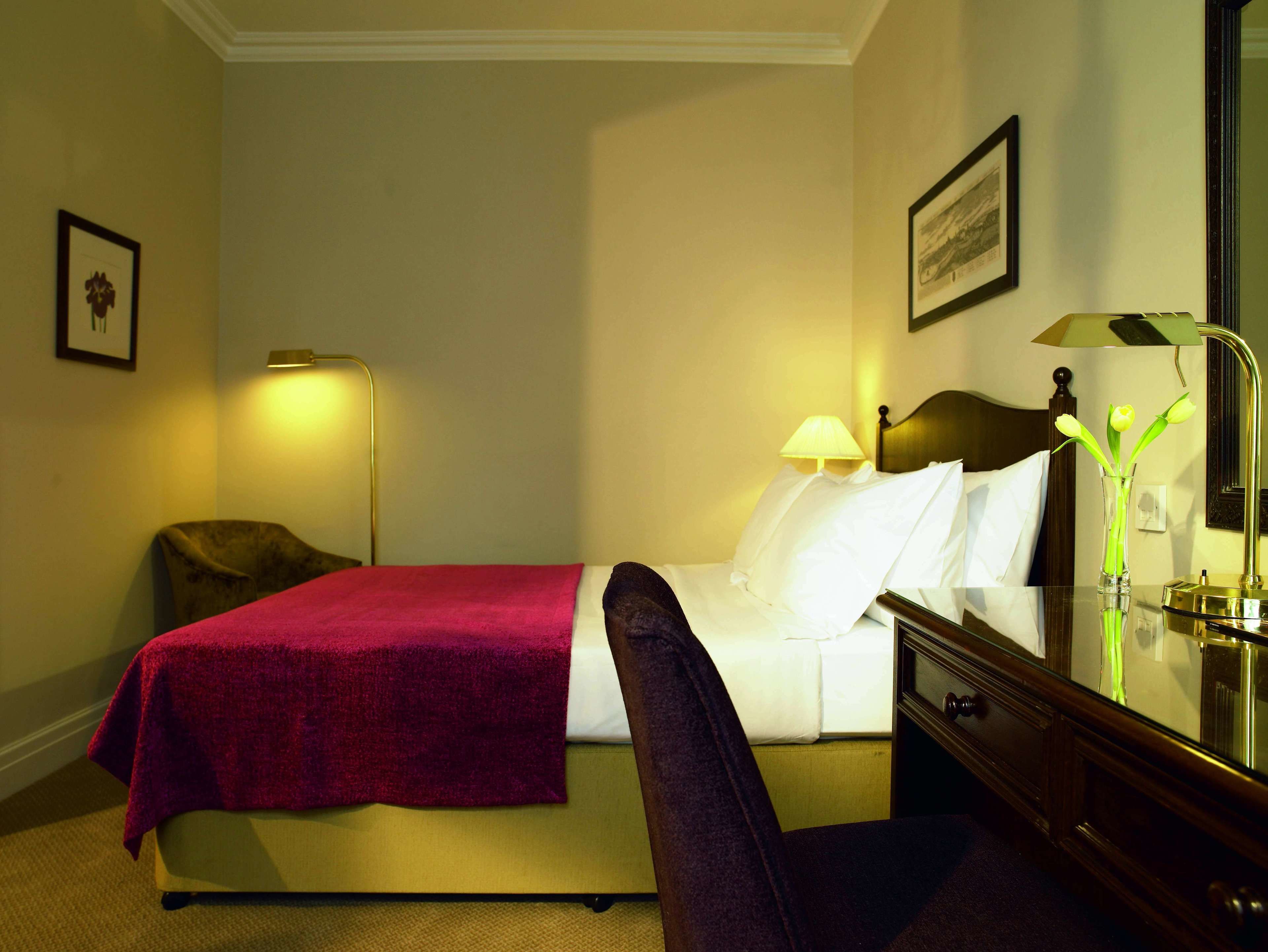 A hotel room with a bed, a chair, a desk with a lamp, and a mirror.