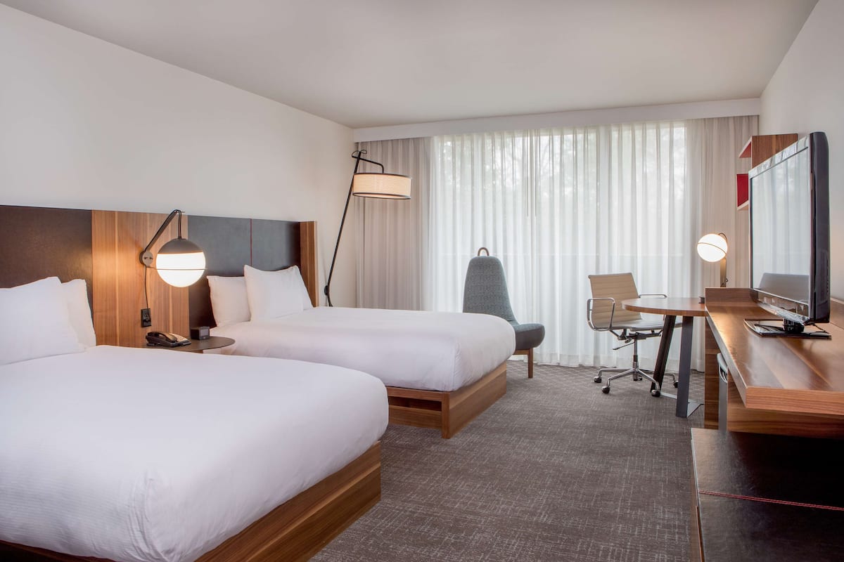A hotel room with two beds, a desk, a chair, a TV, and a large window with sheer curtains.