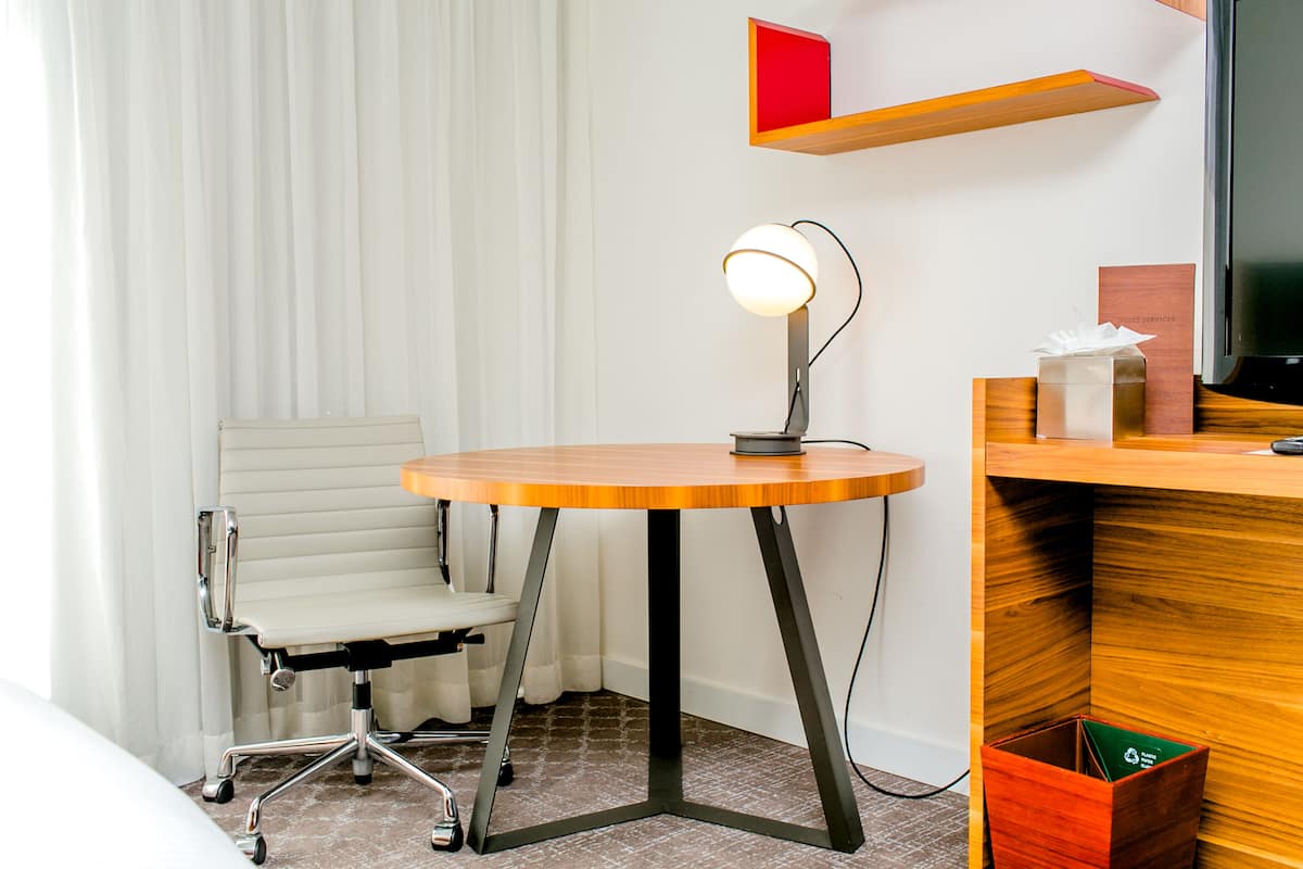 A hotel room with a round wooden table, a white office chair, a desk with a computer monitor, and a small red shelf above the desk.