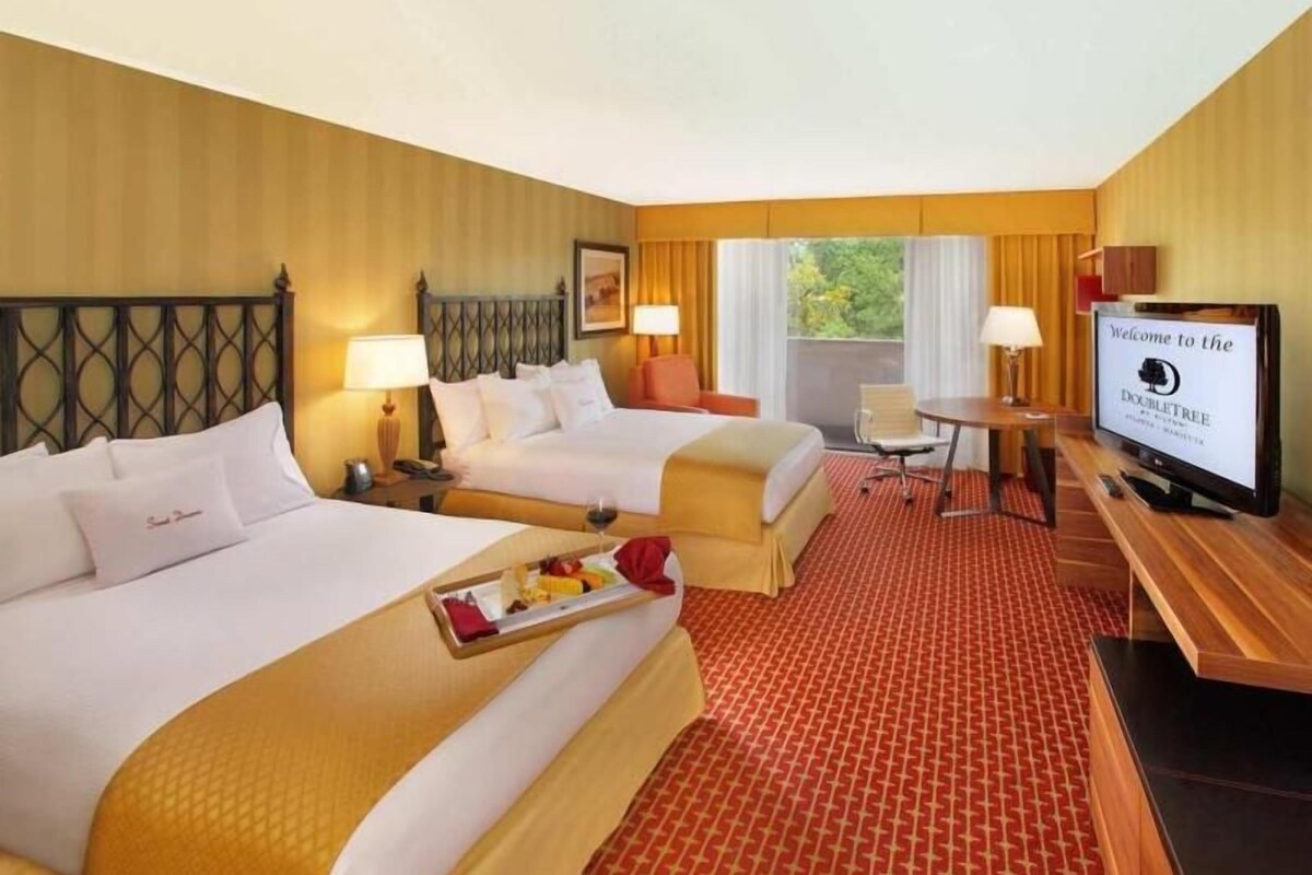 A hotel room with two beds, a TV, a desk, and a window with curtains.