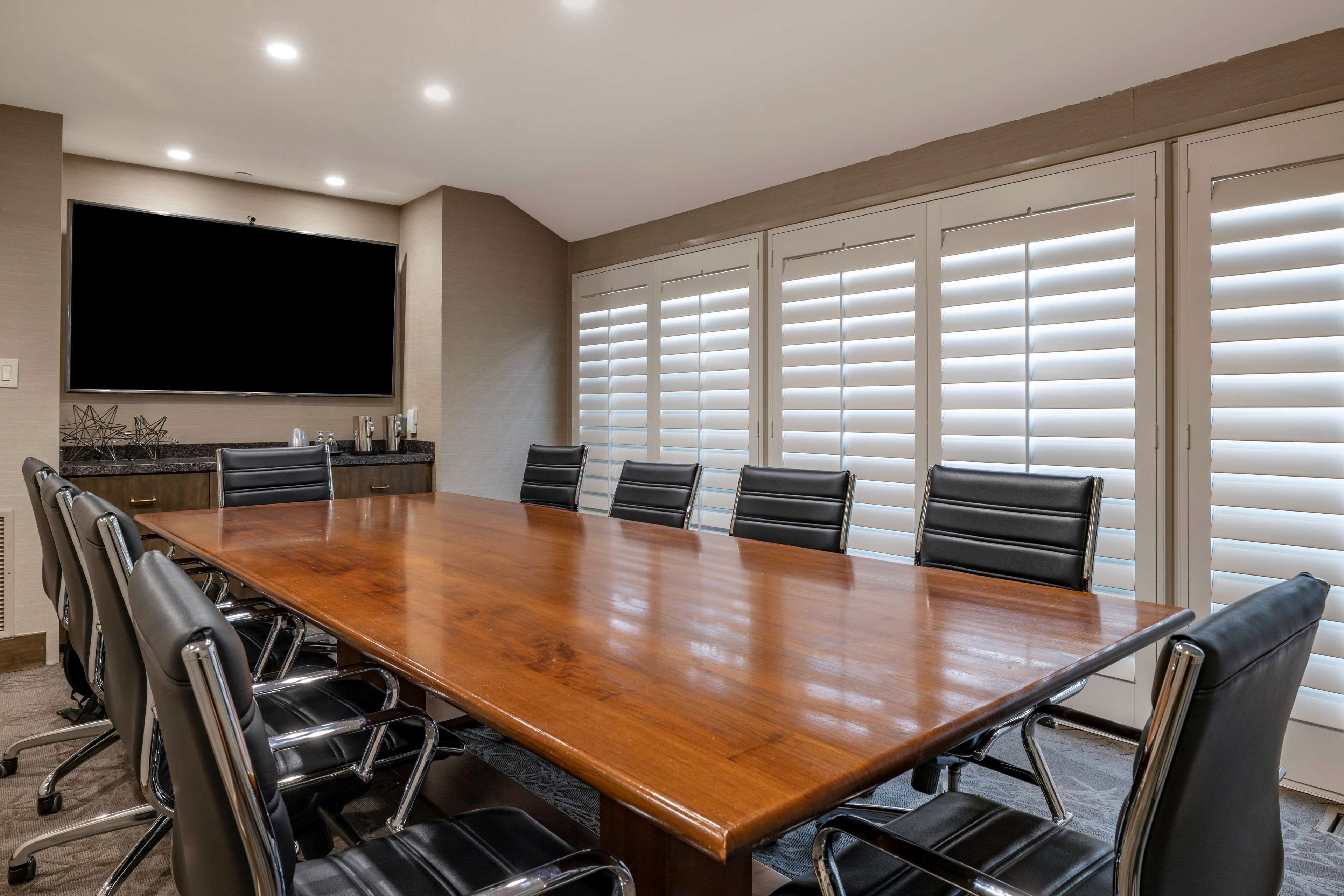 Una sala de conferencias con una amplia mesa de madera, sillas de cuero negro, un televisor de pantalla plana y un ventanal con persianas.