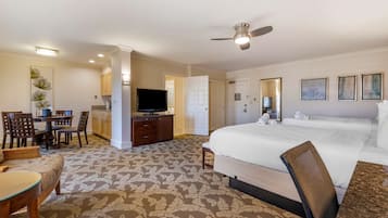 Quarto de hotel com uma cama grande, uma escrivaninha com televisão, área de refeições com cadeiras e piso coberto por carpete.