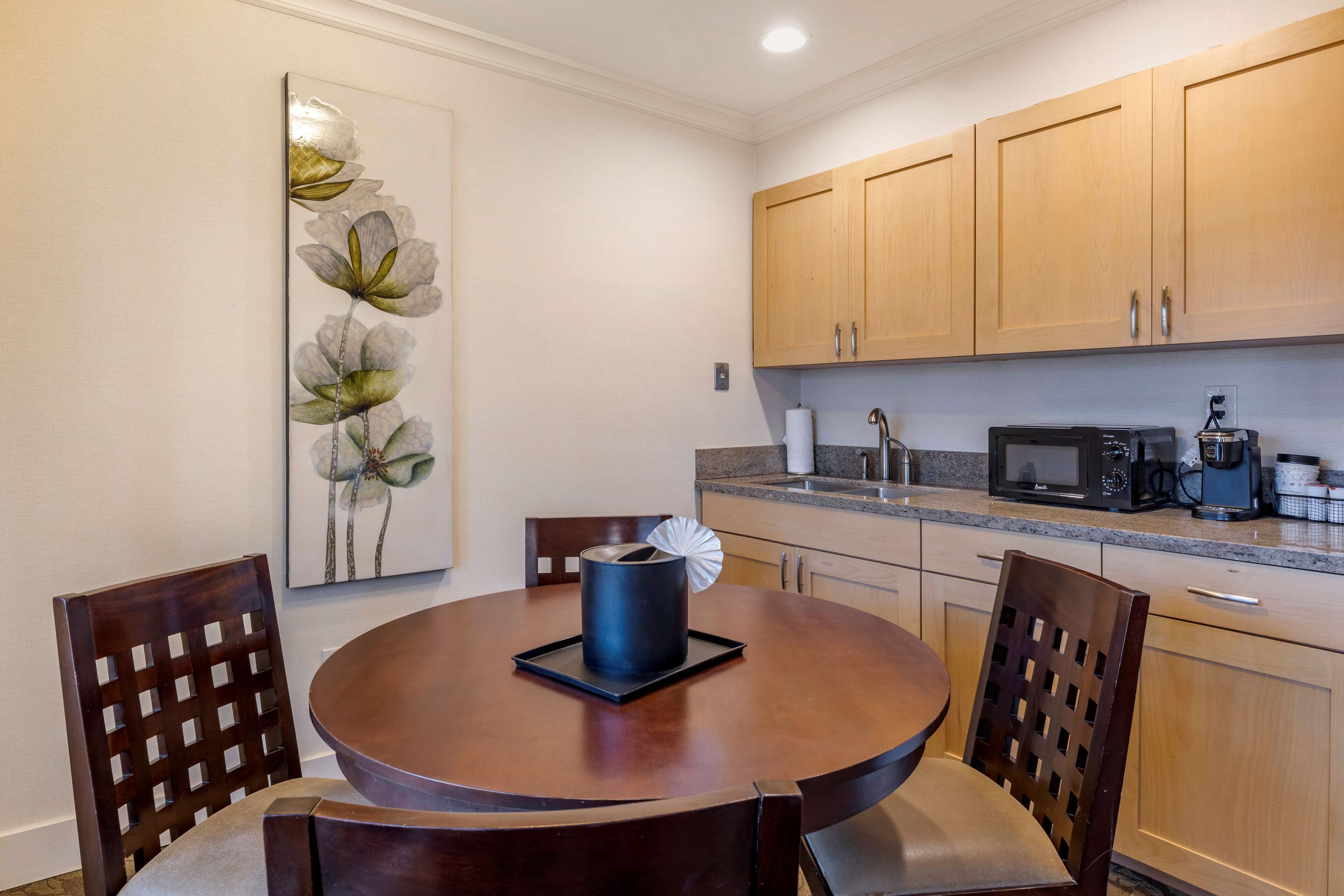 A dining area with a round wooden table, four chairs, a wall-mounted flower painting, wooden cabinets, a microwave, and a coffee maker.