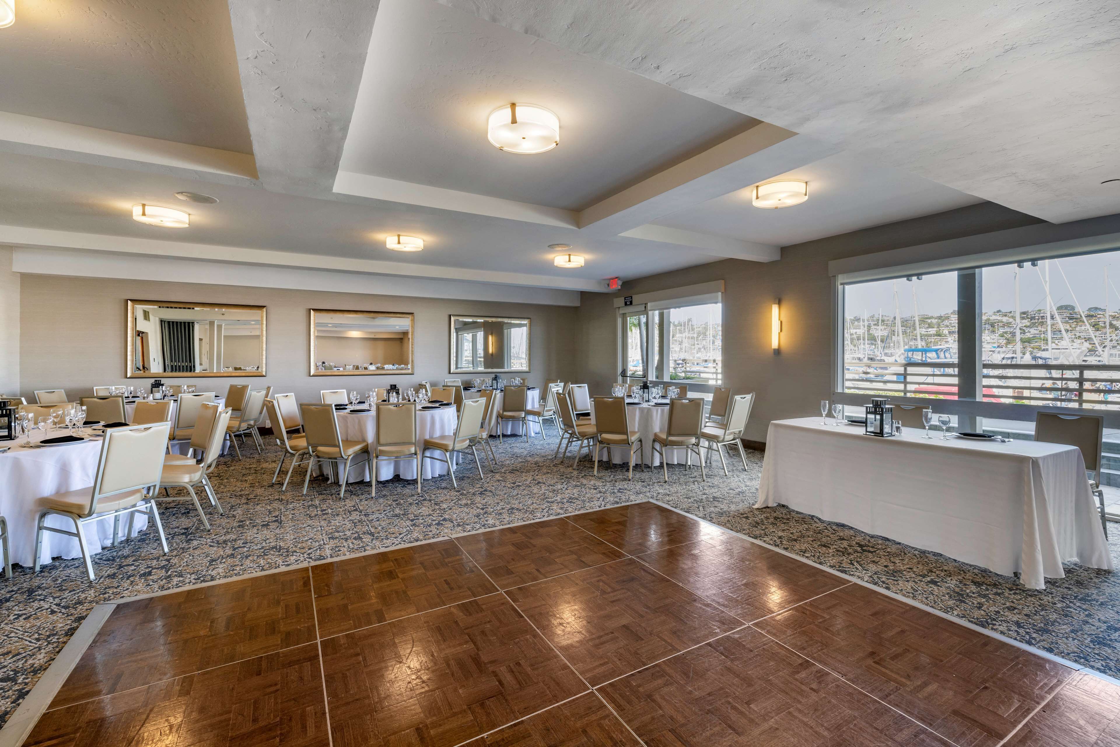 A spacious conference room with multiple tables and chairs, large windows with a view of boats, and a polished wooden dance floor.