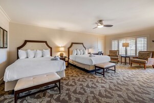 A hotel room with two beds, a sitting area with a chair and a small table, and a large window with blinds.