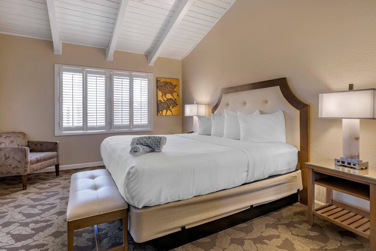 Une chambre d’hôtel avec un grand lit, une chaise, une table de chevet avec une lampe et une fenêtre avec des stores.