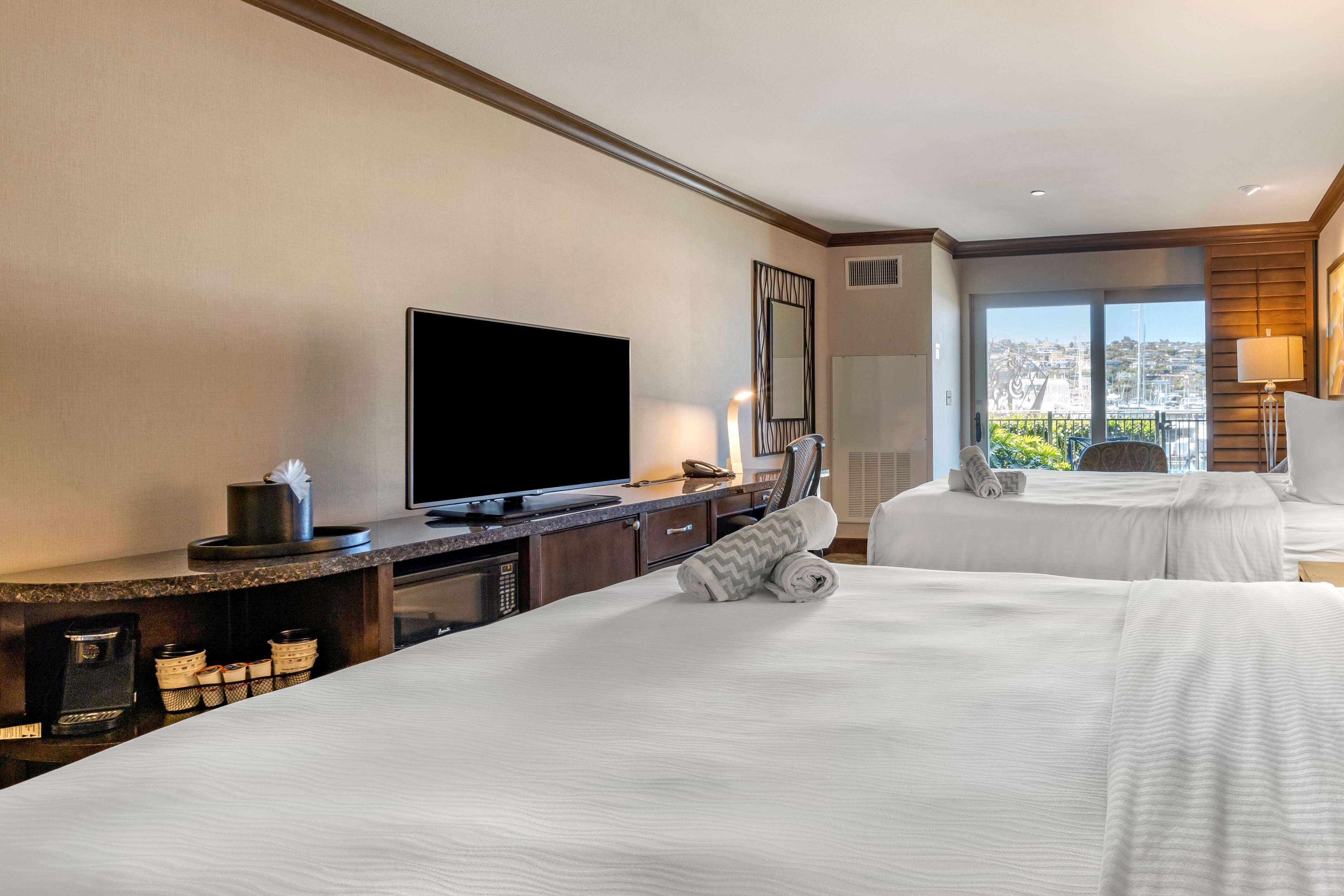 Habitación de hotel con una cama grande, televisión, escritorio y cafetera, y vistas a la ciudad a través de amplios ventanales.