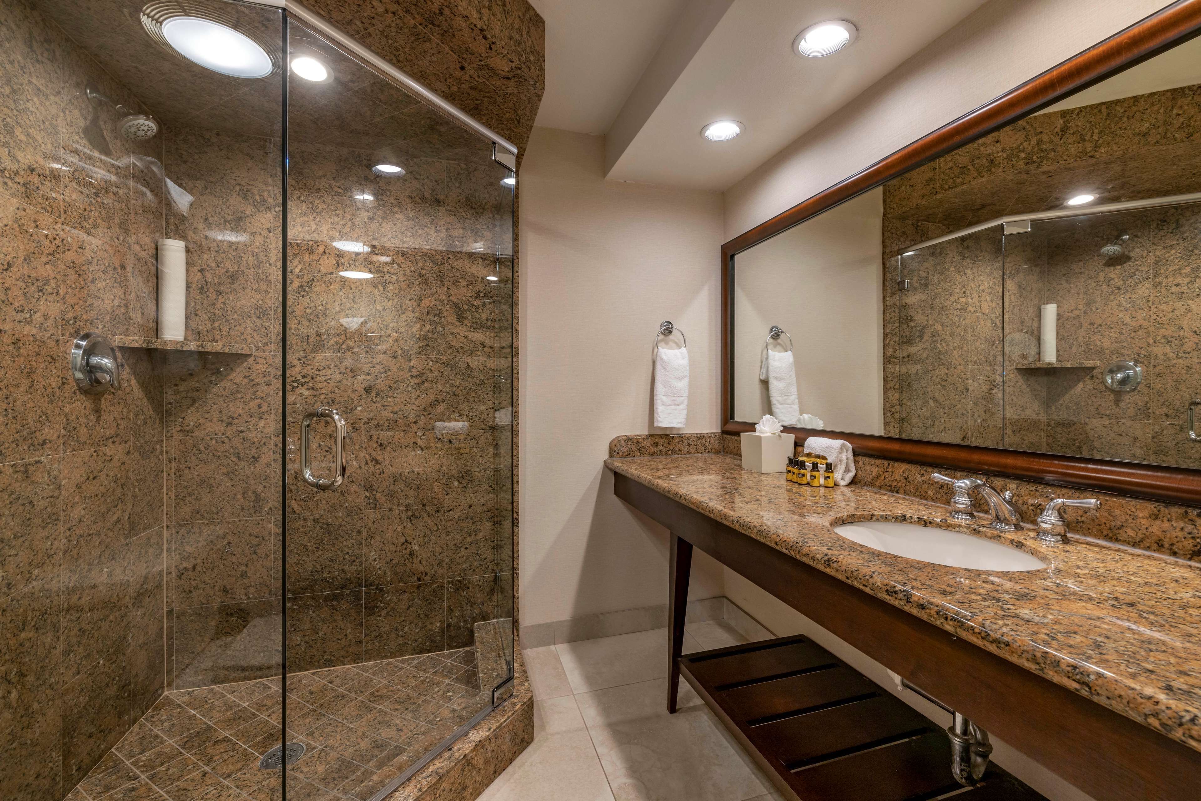 Un baño moderno con cabina de ducha de vidrio, encimeras de granito, un espejo grande y un mueble de tocador de madera.
