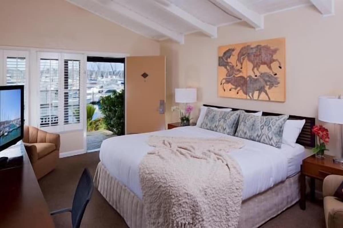 A hotel room with a large bed, a television, a desk, and a balcony with a view of the marina.