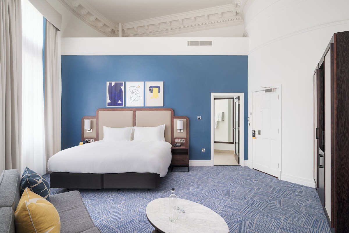 Habitación de hotel con una cama grande, una mesita de noche, una lámpara, un espejo y vistas al pasillo.