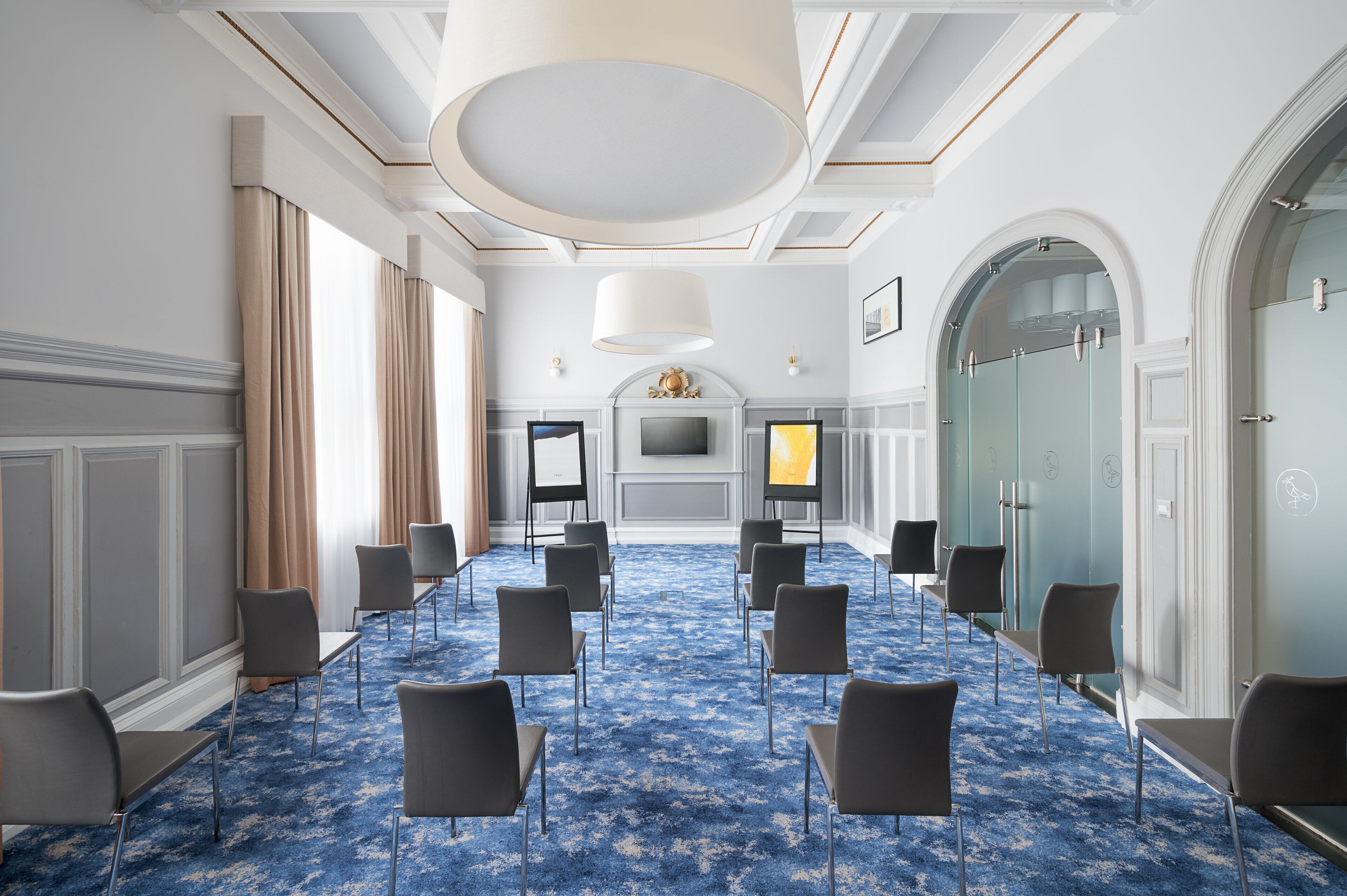 A conference room with a blue carpet, white walls, and a large chandelier. There are rows of chairs and a TV screen.