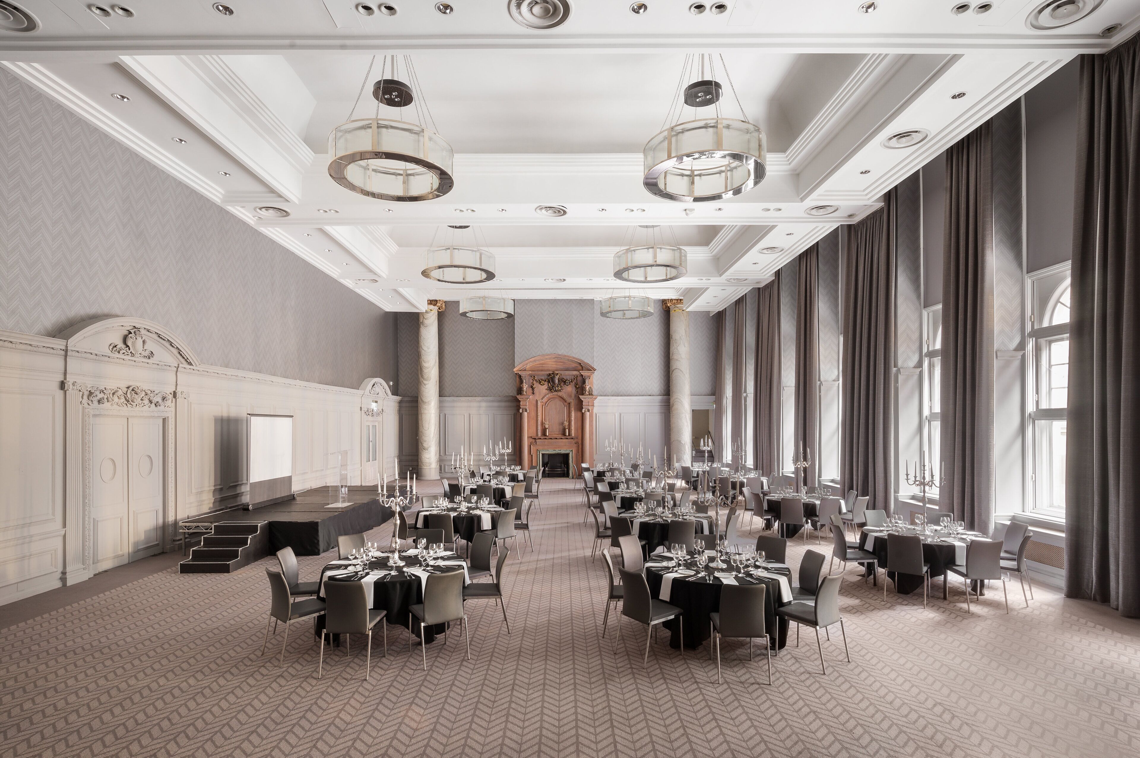 A large banquet hall with multiple round tables, chandeliers, and a fireplace.