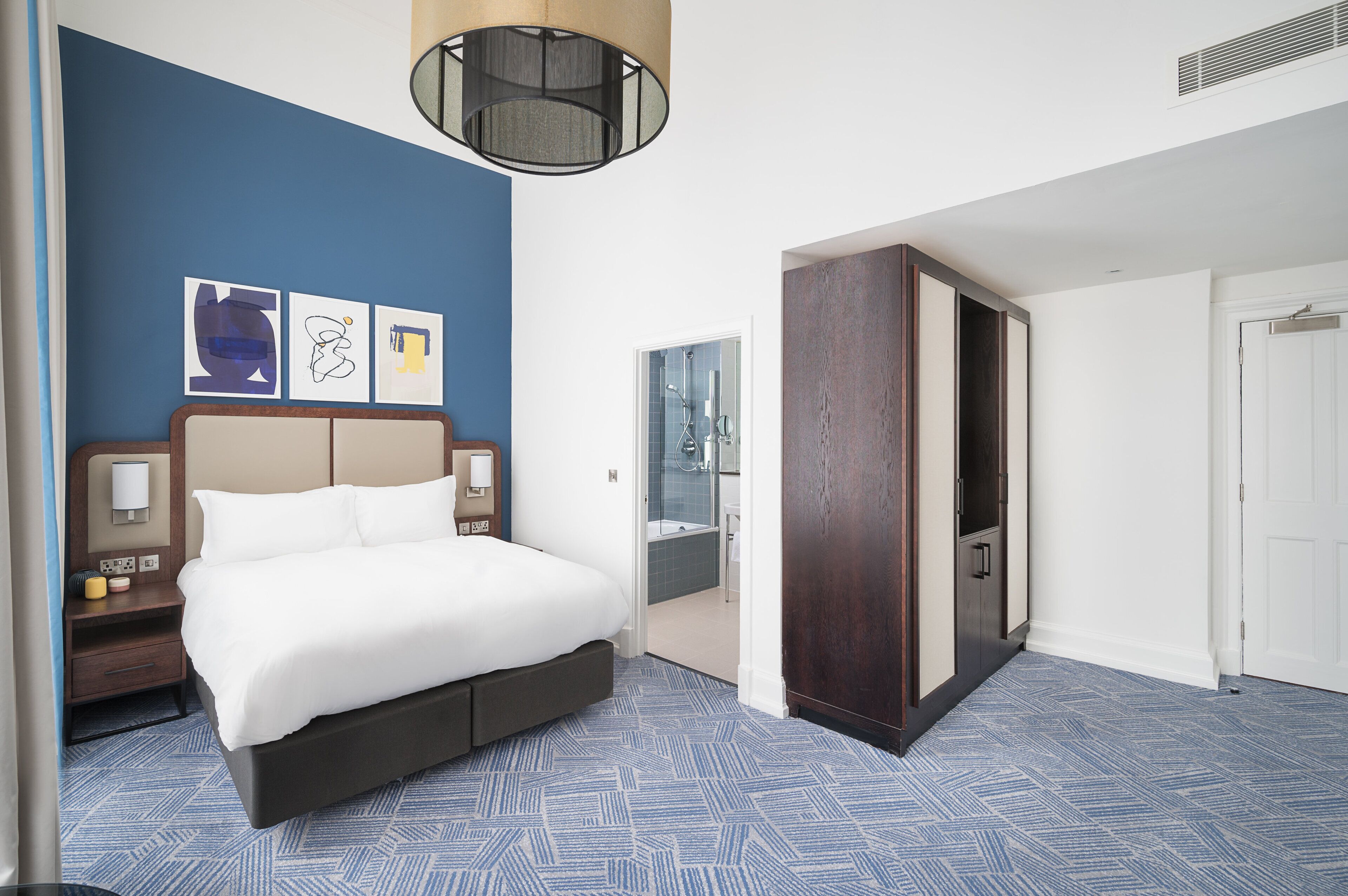 Une chambre d’hôtel avec un grand lit, une armoire, une table de chevet avec une lampe et des œuvres d’art accrochées au mur.