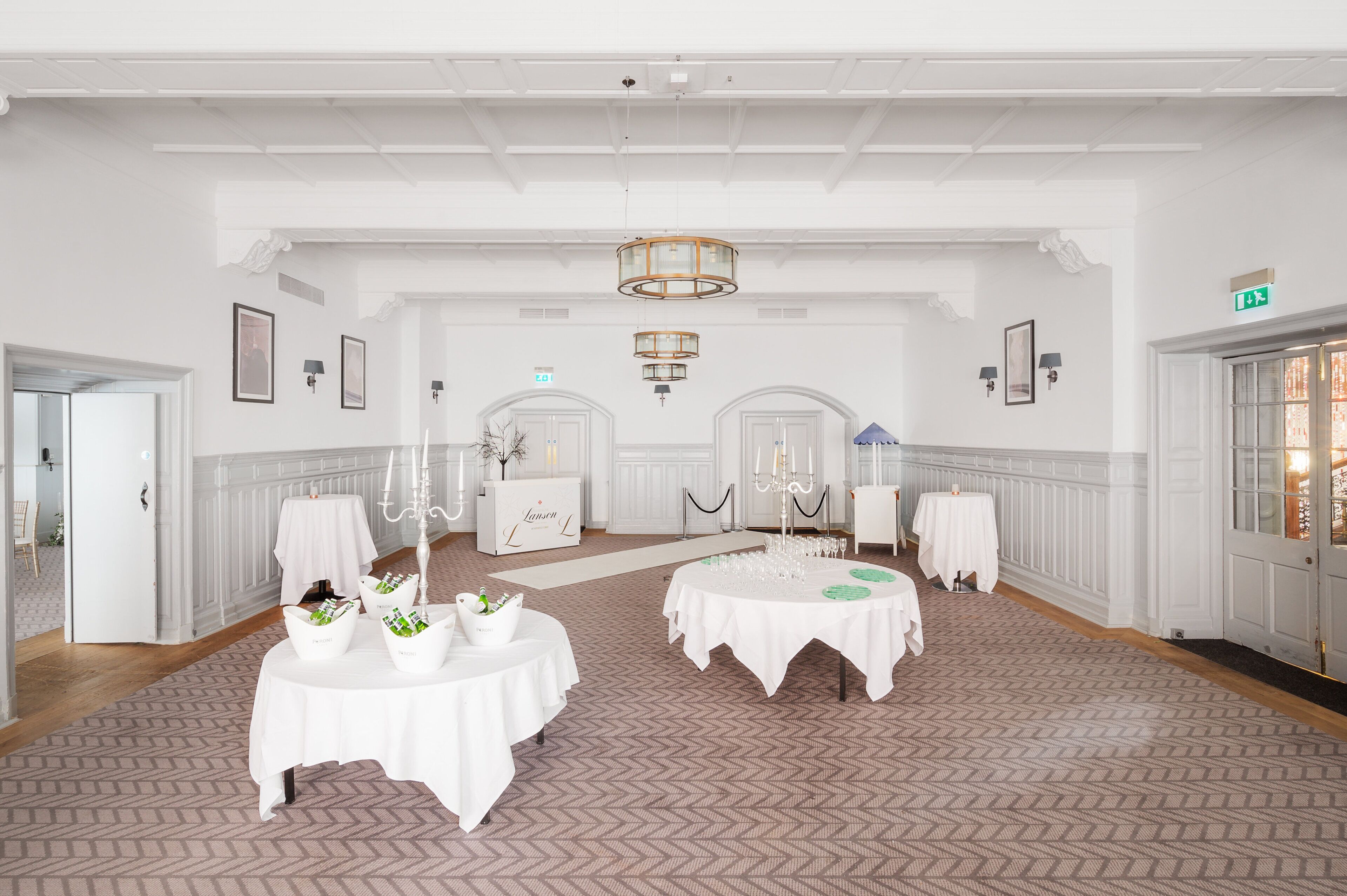A spacious hall with high ceilings and chandeliers, featuring multiple round tables with white tablecloths and centerpieces, and a patterned carpet.