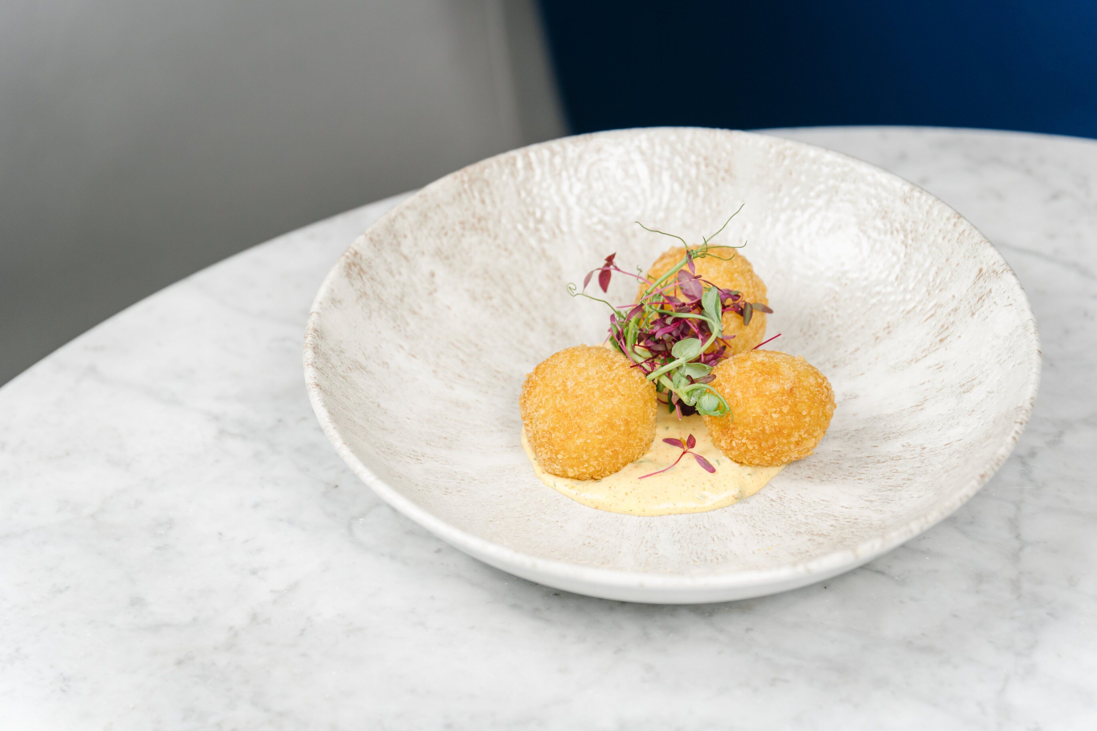 A white ceramic plate with three round, golden-brown items and a creamy sauce, garnished with microgreens, on a marble surface.
