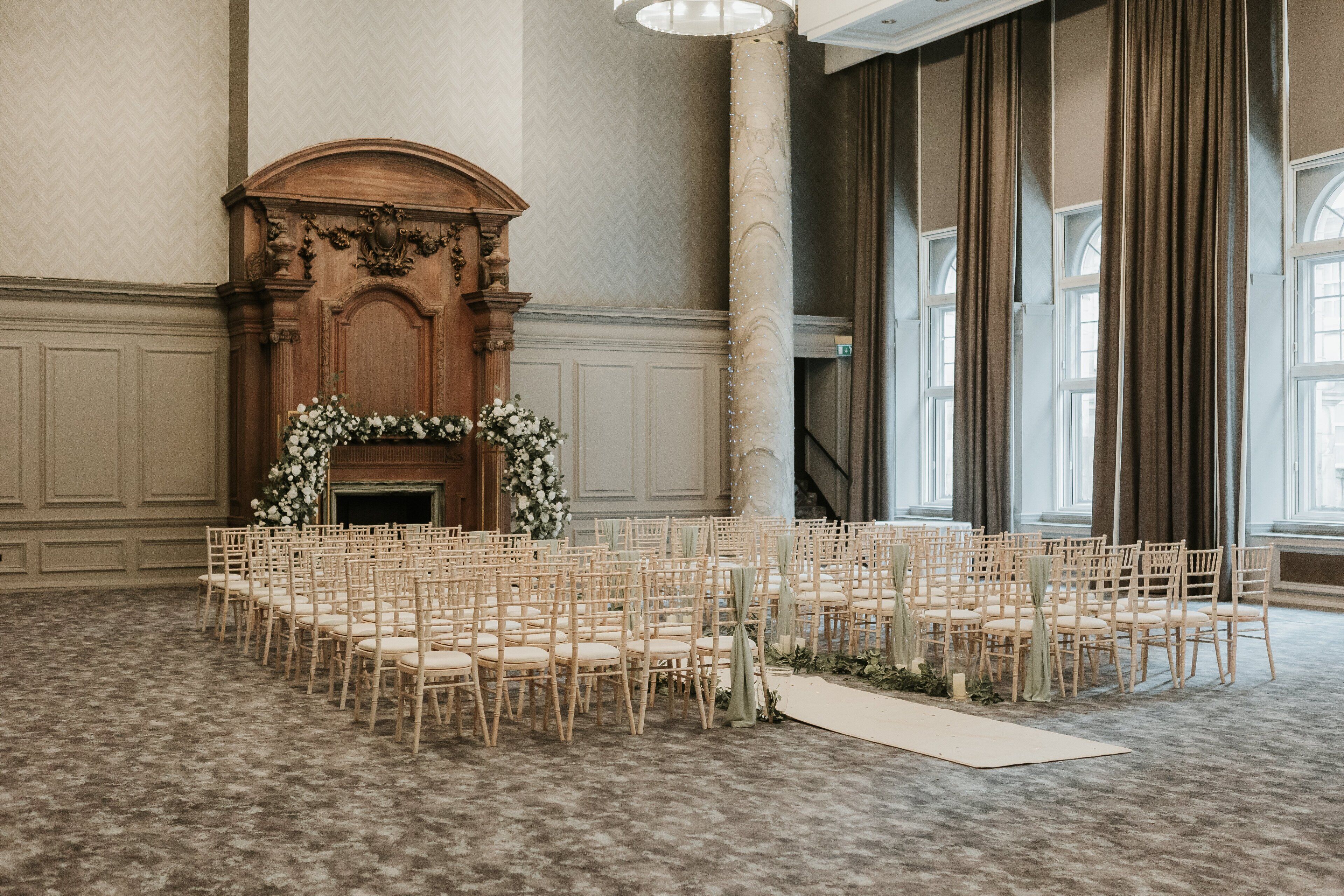 A formal event space with rows of wooden chairs arranged for seating, a fireplace with a floral arrangement, and large windows with curtains.