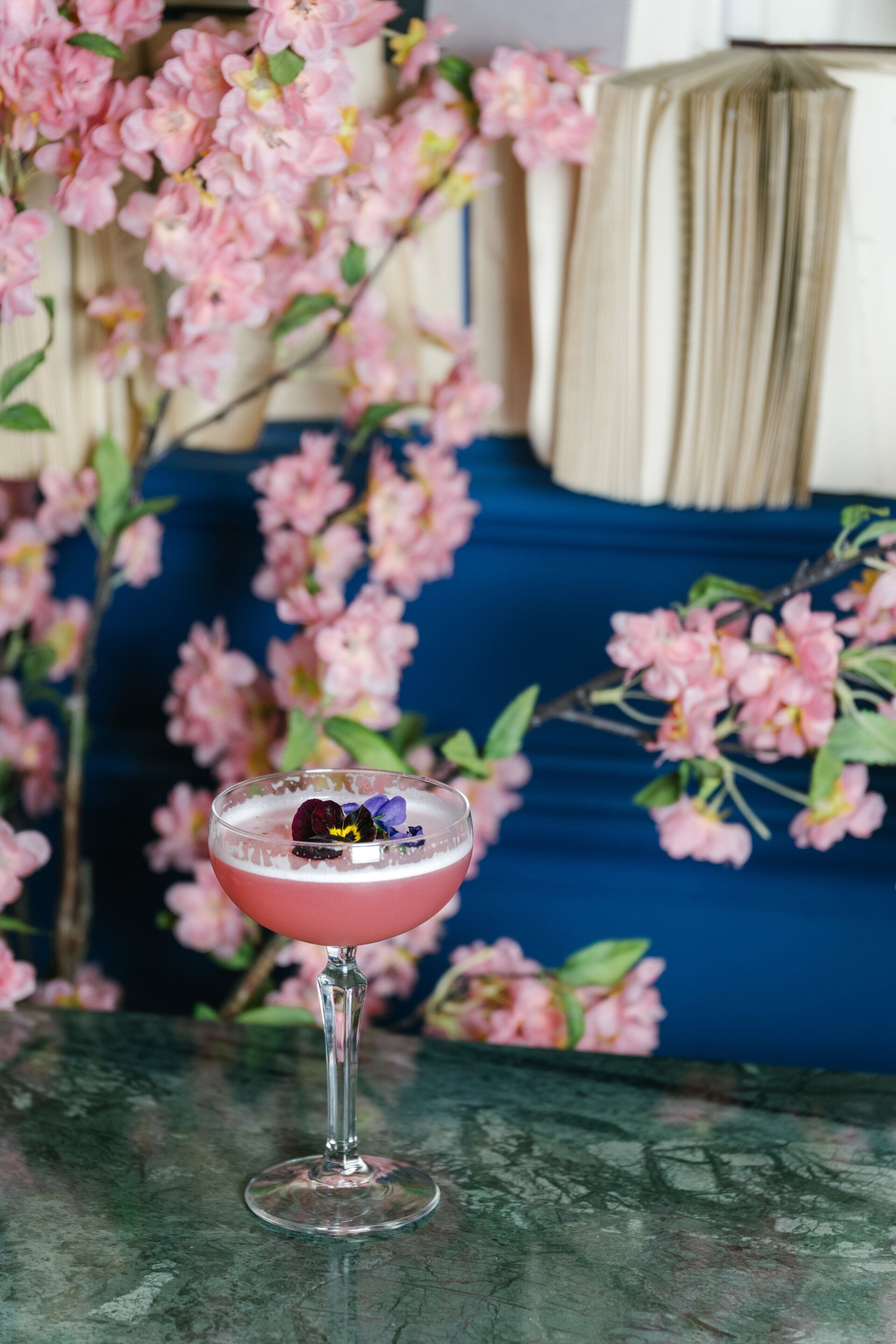 A cocktail in a glass on a table with a blue sofa and pink flowers in the background.