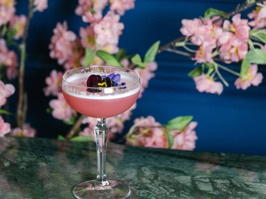 Un cóctel en un vaso sobre una mesa, con un sofá azul y flores rosadas al fondo.