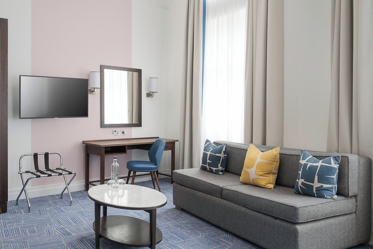 Una habitación de hotel moderna con televisor de pantalla plana, escritorio, silla azul, sofá gris con cojines estampados y una mesita redonda.