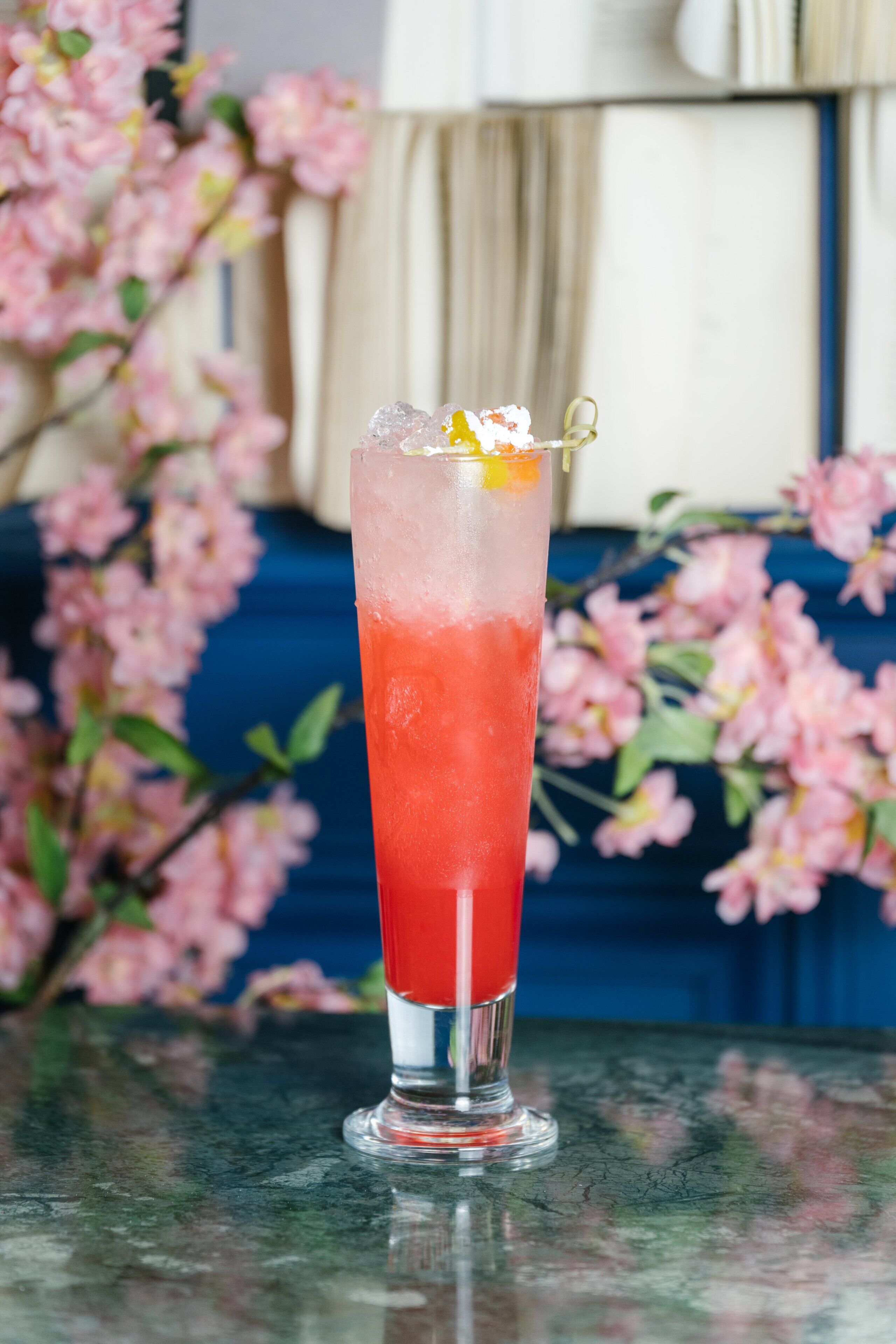A tall glass with a layered drink, garnished with a fruit and a decorative item, set against a backdrop of pink flowers and a blue wall.