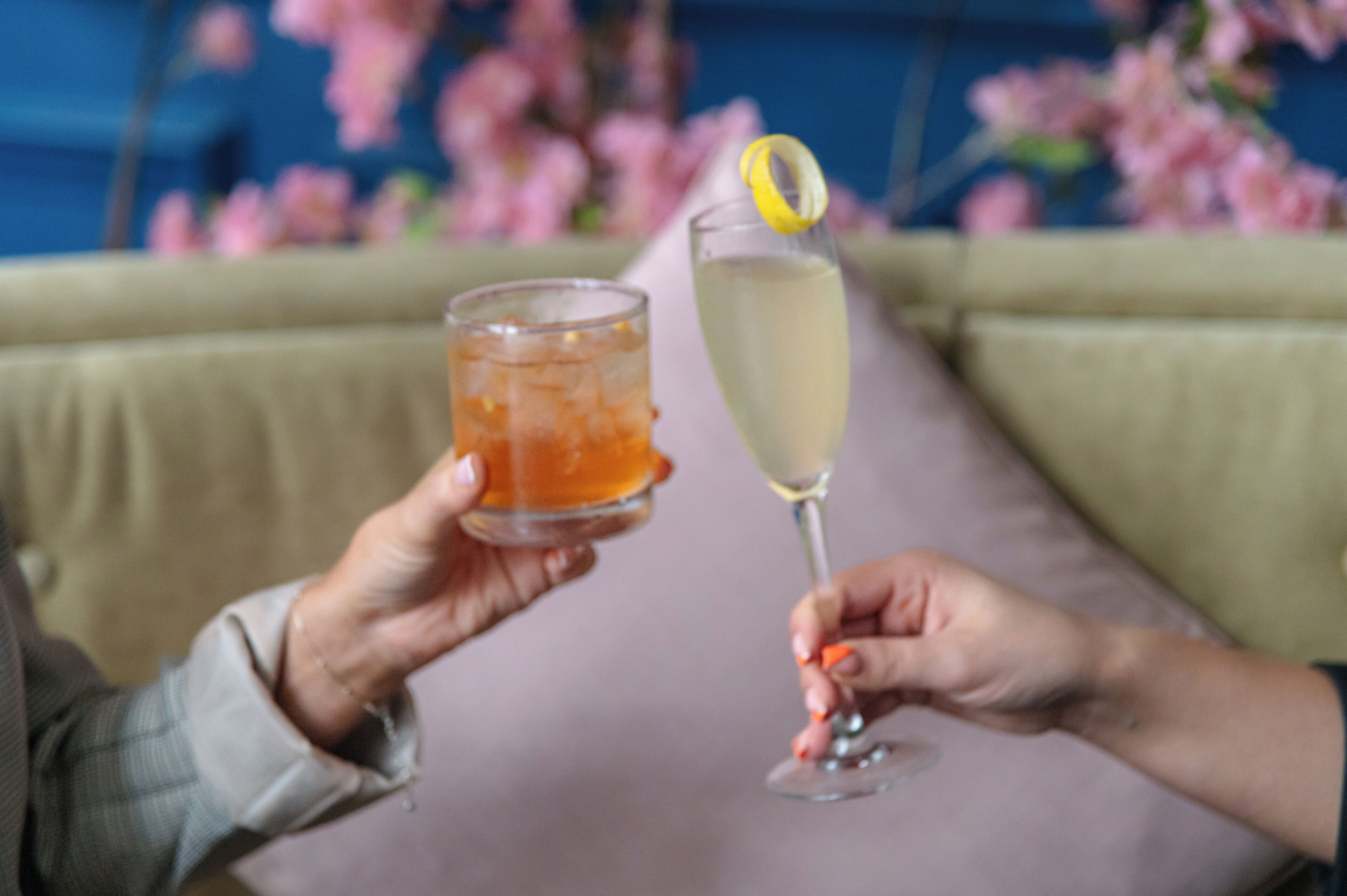 Dos manos sosteniendo bebidas, una con un vaso y la otra con una copa de champán, sobre una mesa con un fondo floral.