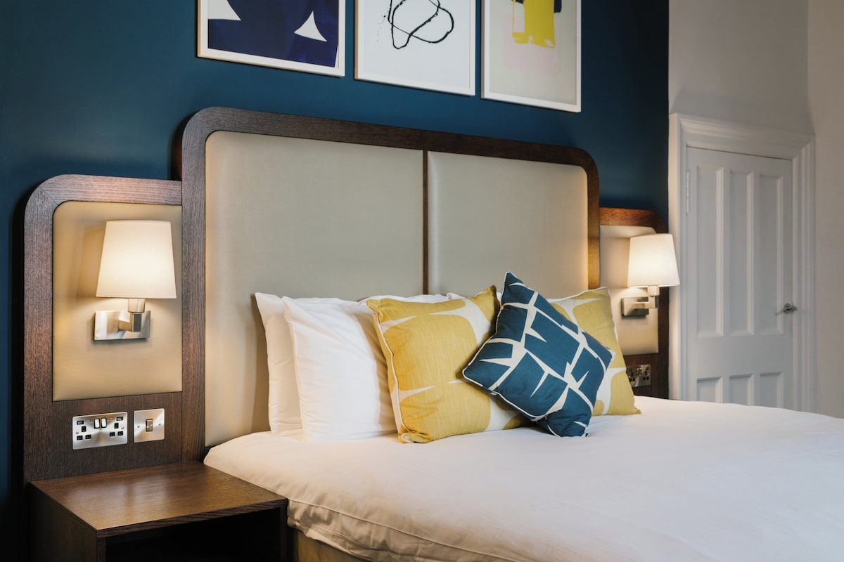 Una cama bien tendida con un cabecero, dos lámparas de noche y una pared decorada con cuadros.