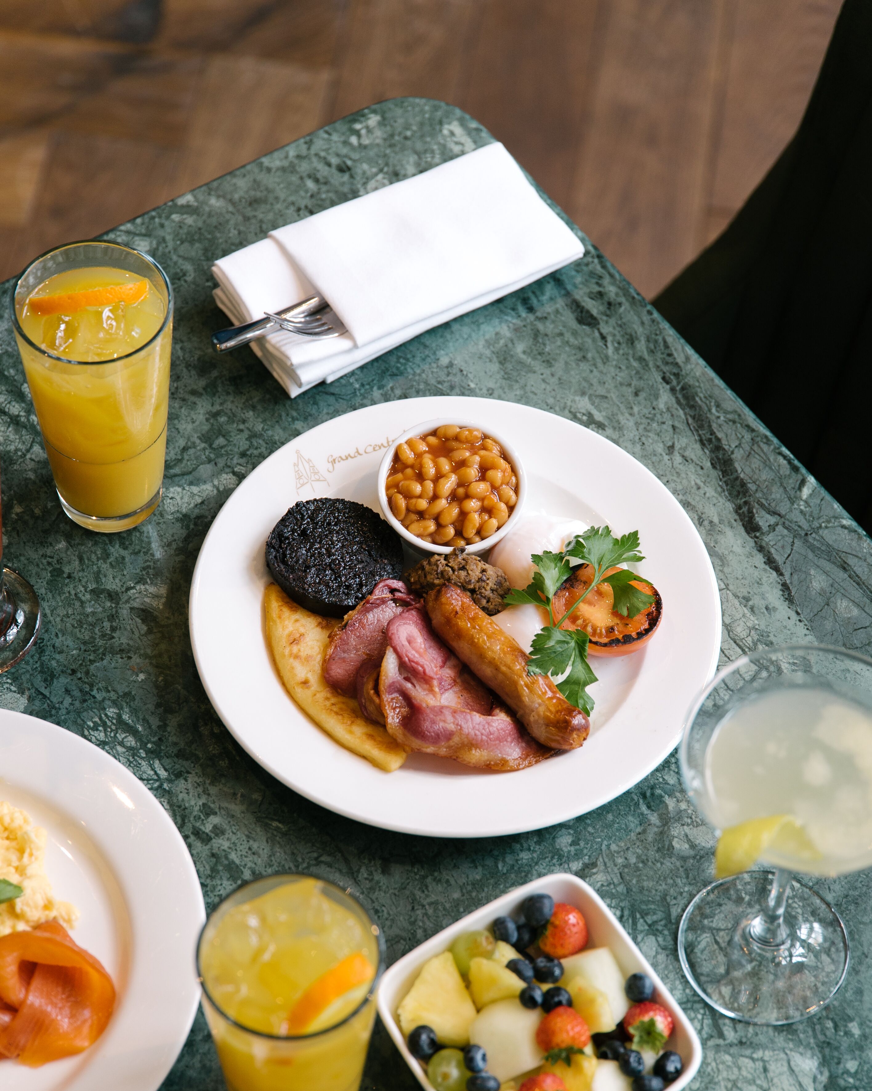A breakfast setting with a plate of eggs, sausages, beans, and a grilled tomato, accompanied by a glass of orange juice and a fruit bowl.