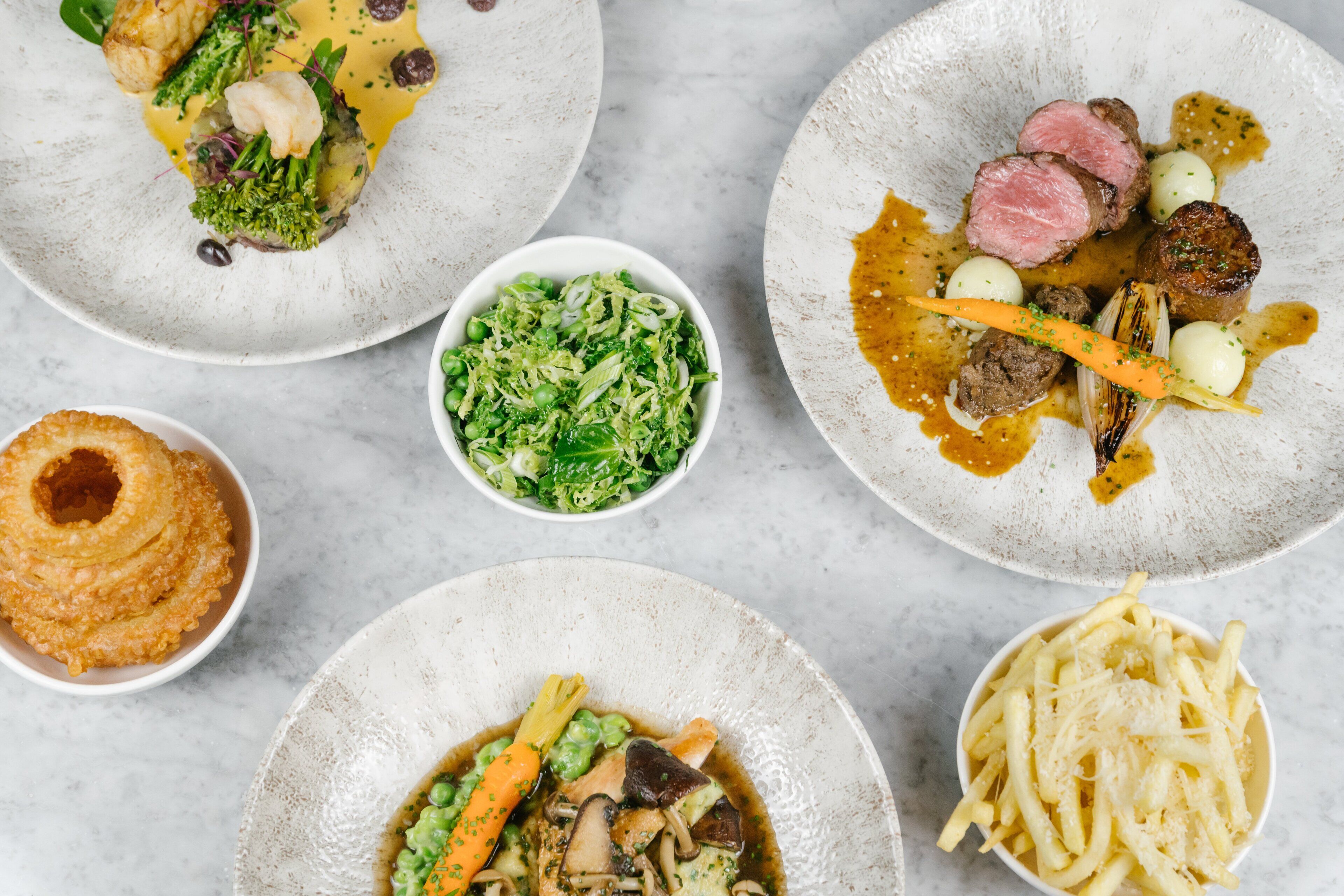 A variety of dishes on plates, including a steak, vegetables, and a bowl of fries.