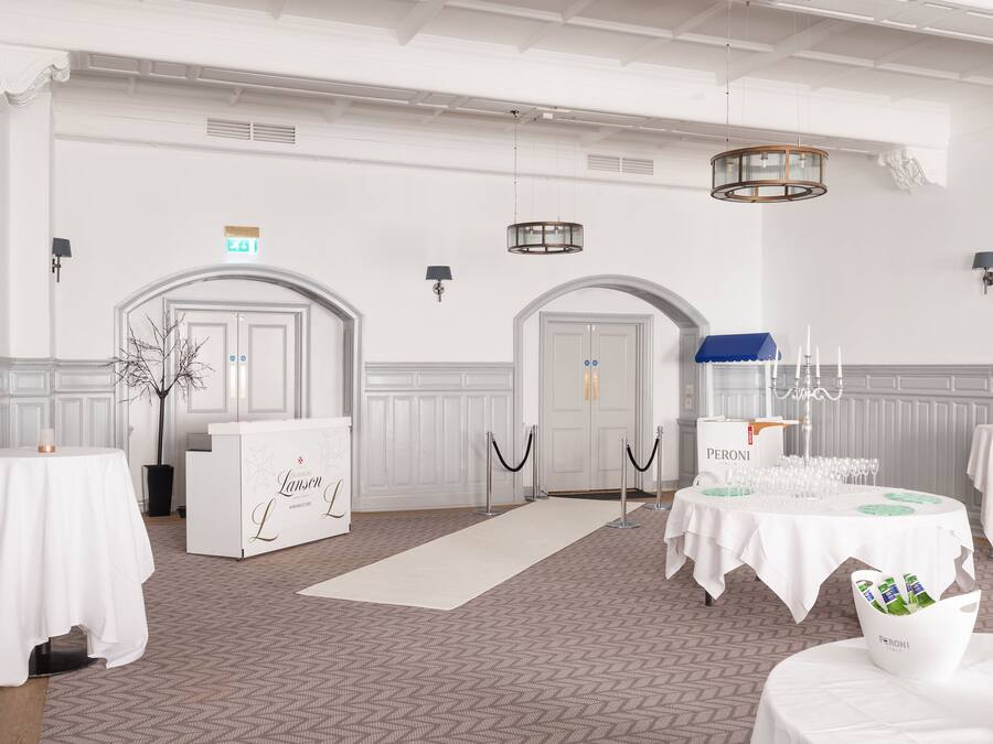 A spacious hall with white walls, arched doorways, and a patterned floor. Tables are set with white tablecloths and wine glasses. A reception desk labeled "Lanson L" is visible.