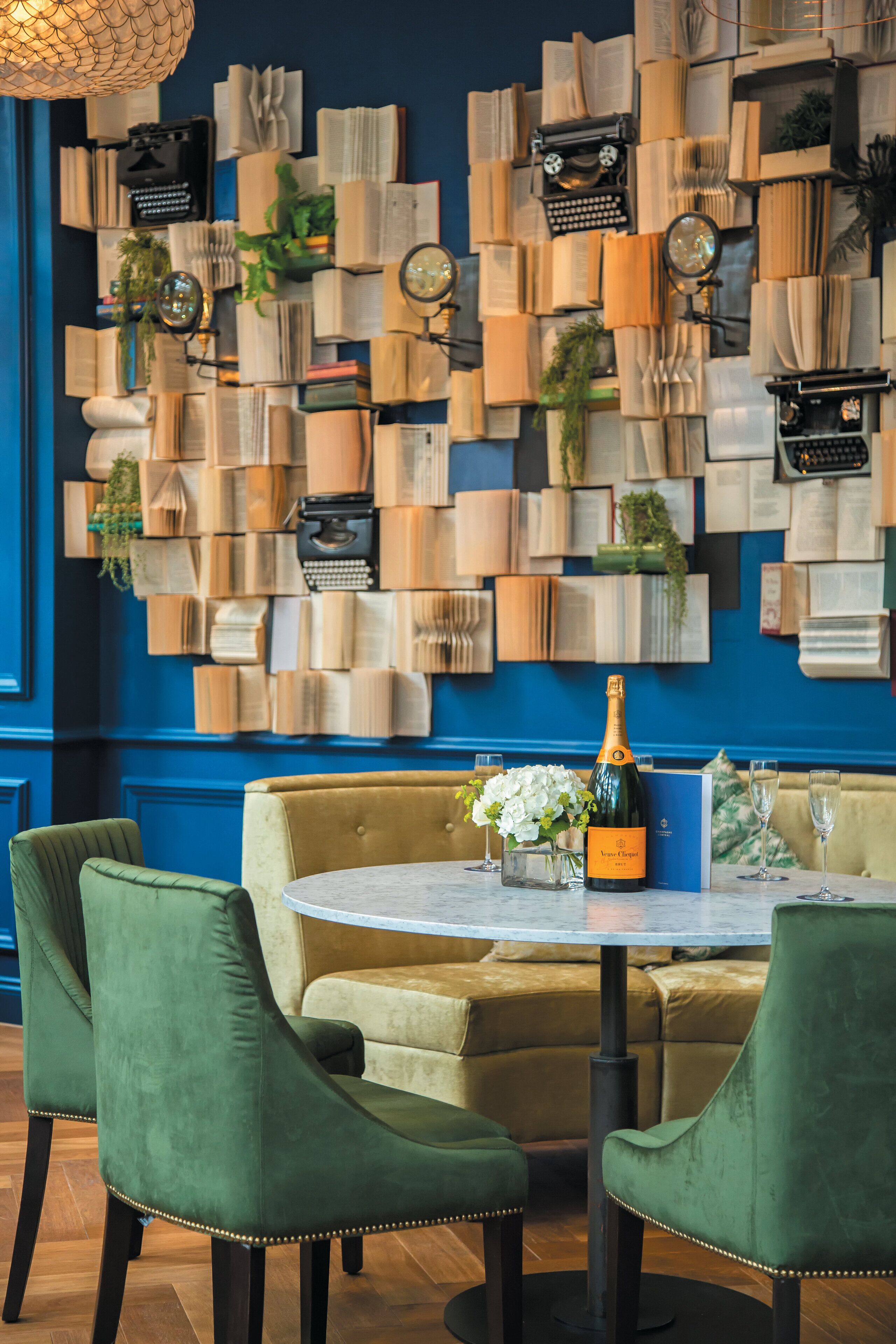 A dining area with a round table, green upholstered chairs, and a wall decorated with books and mirrors.