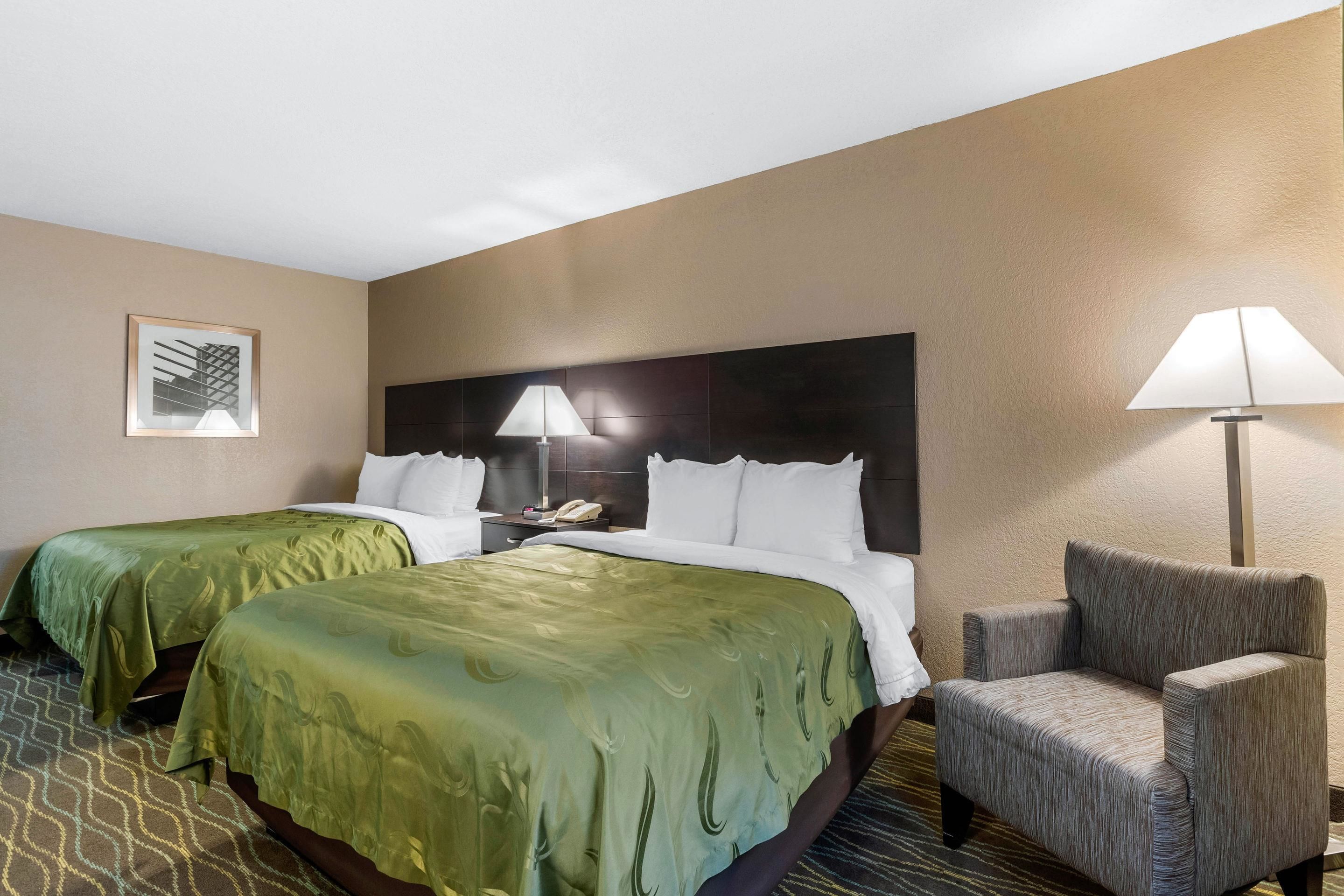 A hotel room with two beds, a nightstand, a lamp, and a chair.