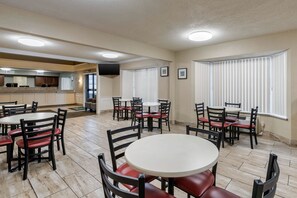 A spacious dining area with multiple round tables and chairs, a kitchen in the background, and large windows with blinds.