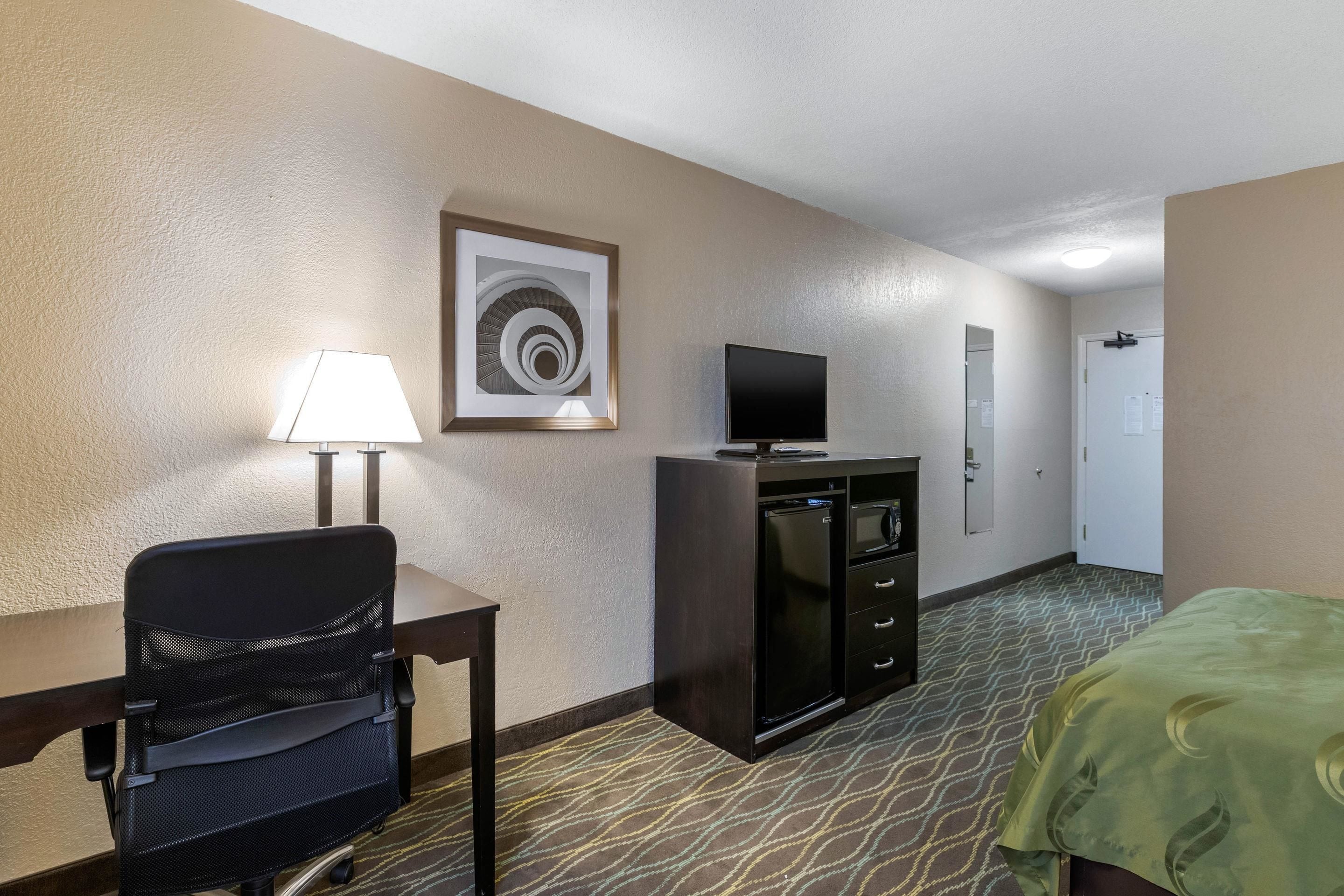 A hotel room with a bed, a desk with a chair, a television, a minibar, and a door.