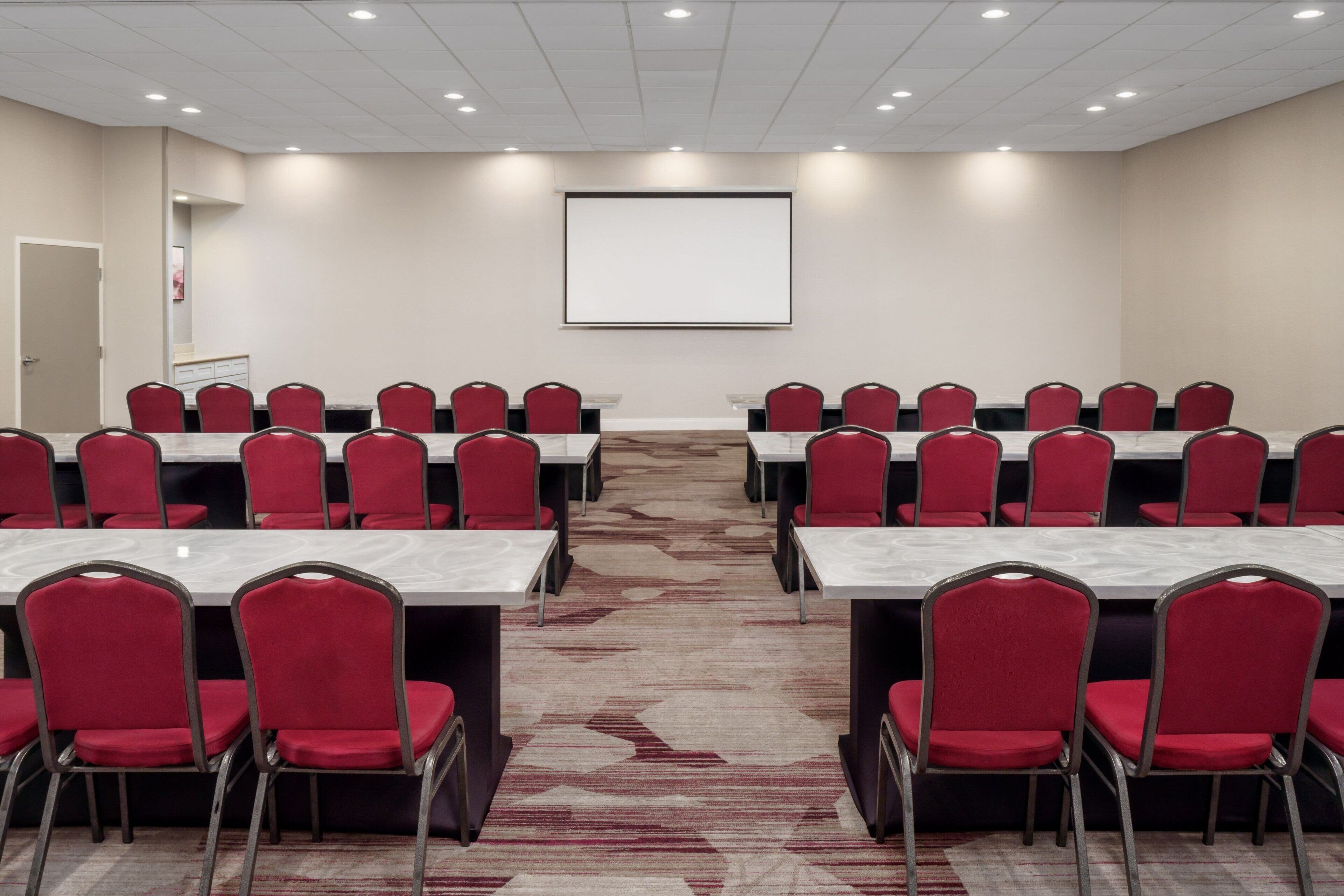 A conference room with rows of red chairs and white tables, a projector screen, and a carpeted floor.