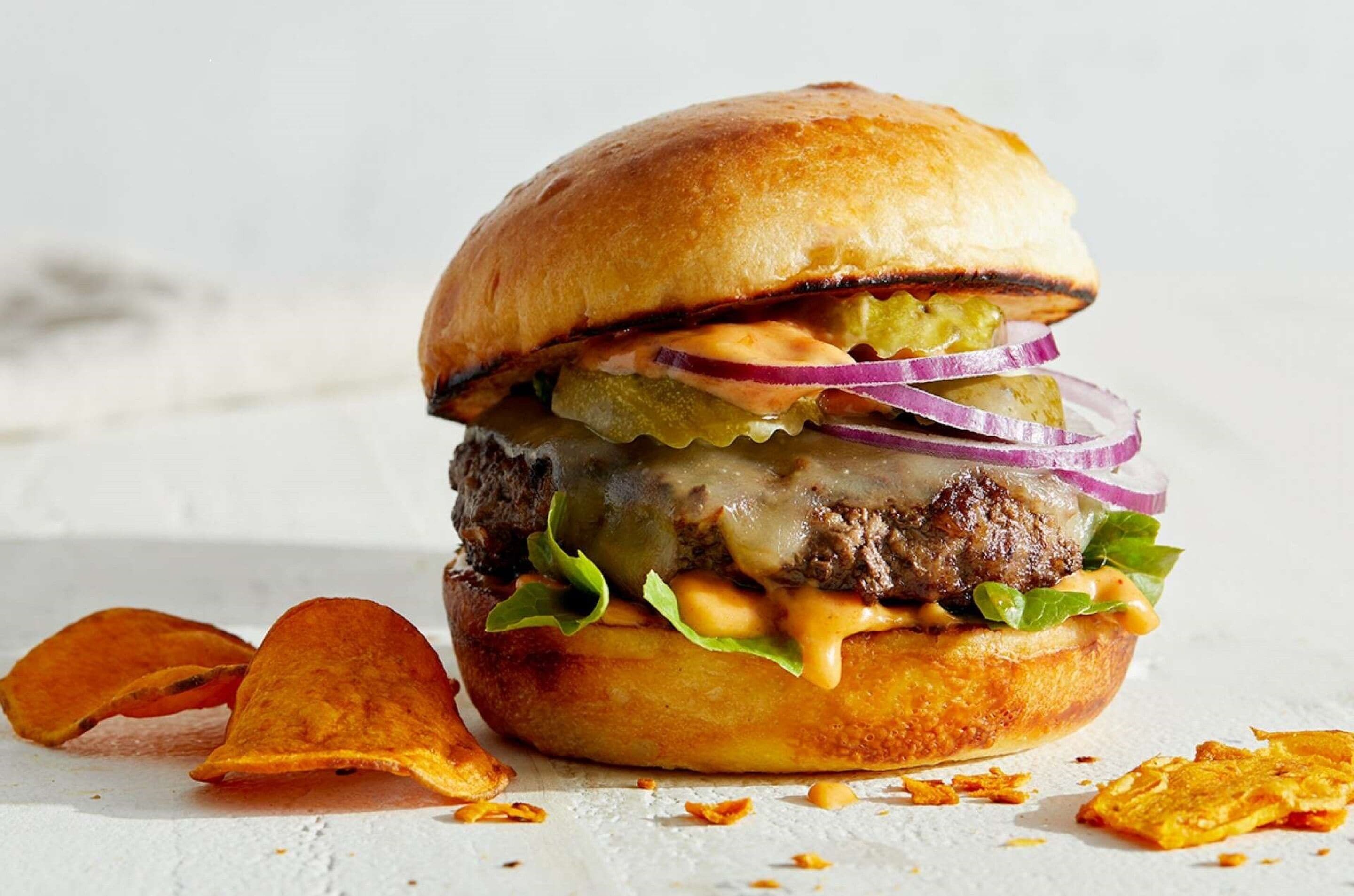 Ein Burger mit Salat, Käse und Gewürzgurken auf einem Brötchen, dazu Chips.