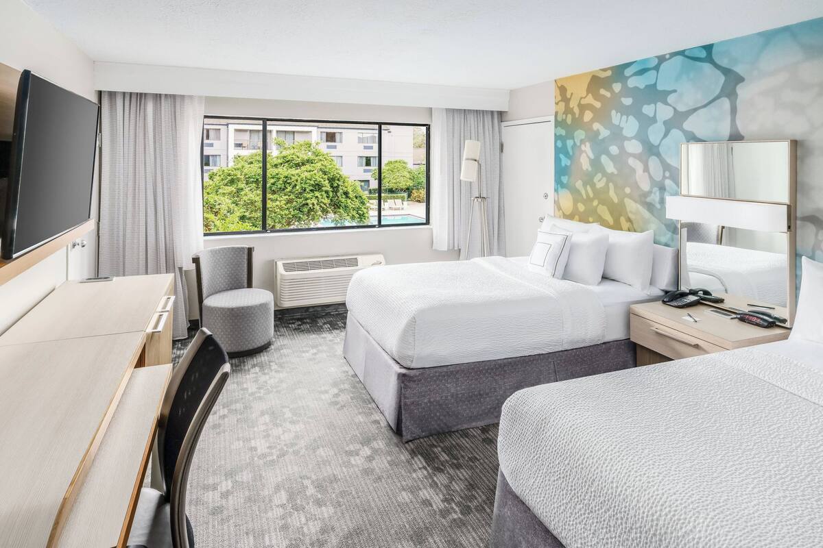 Camera d'albergo con un letto, una scrivania, una sedia, una TV e una finestra con vista su edifici e alberi.