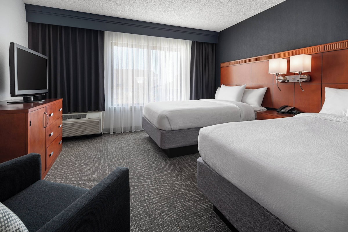 A hotel room with two beds, a TV, a desk with a lamp, and a large window with curtains.