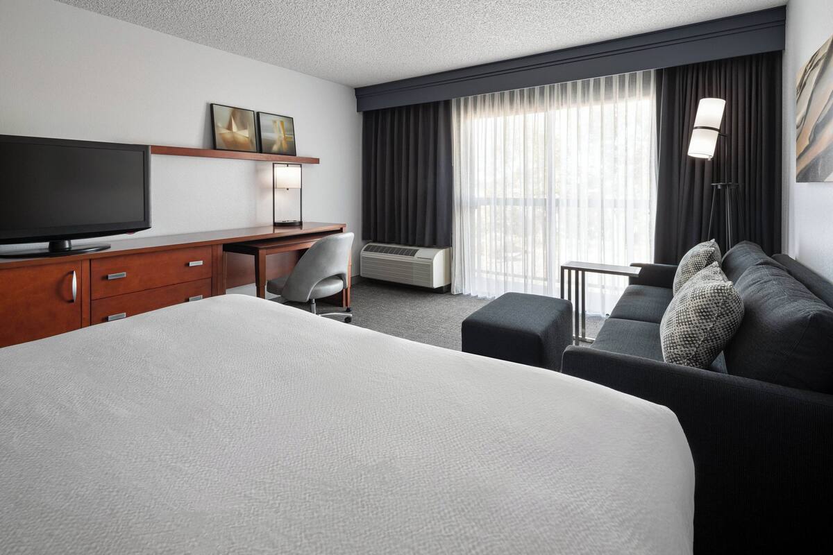 A hotel room with a large bed, a desk with a flat-screen TV, a chair, a sofa, and a window with curtains.