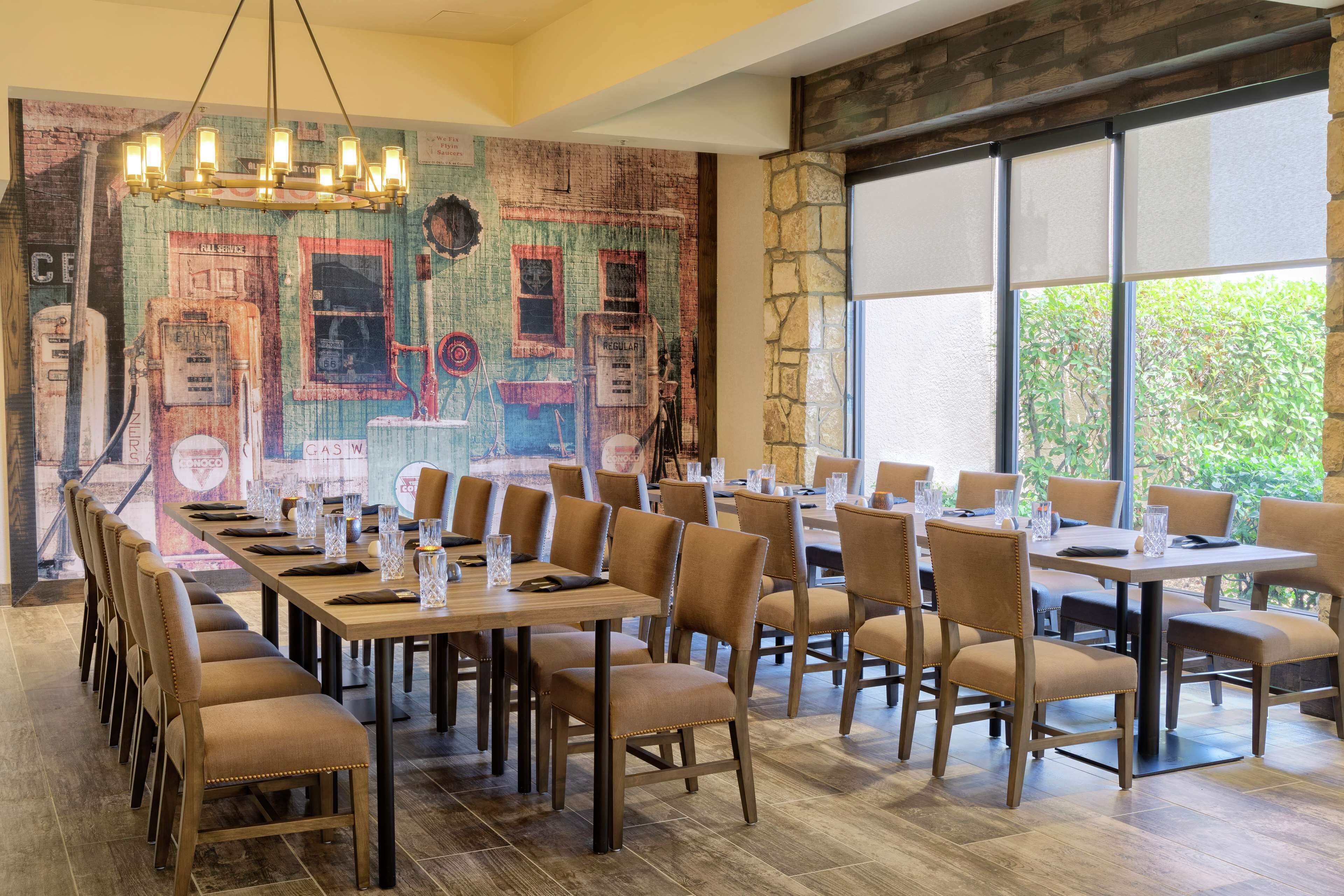 A dining area with long tables and chairs, a mural on the wall, and large windows with a view of greenery.
