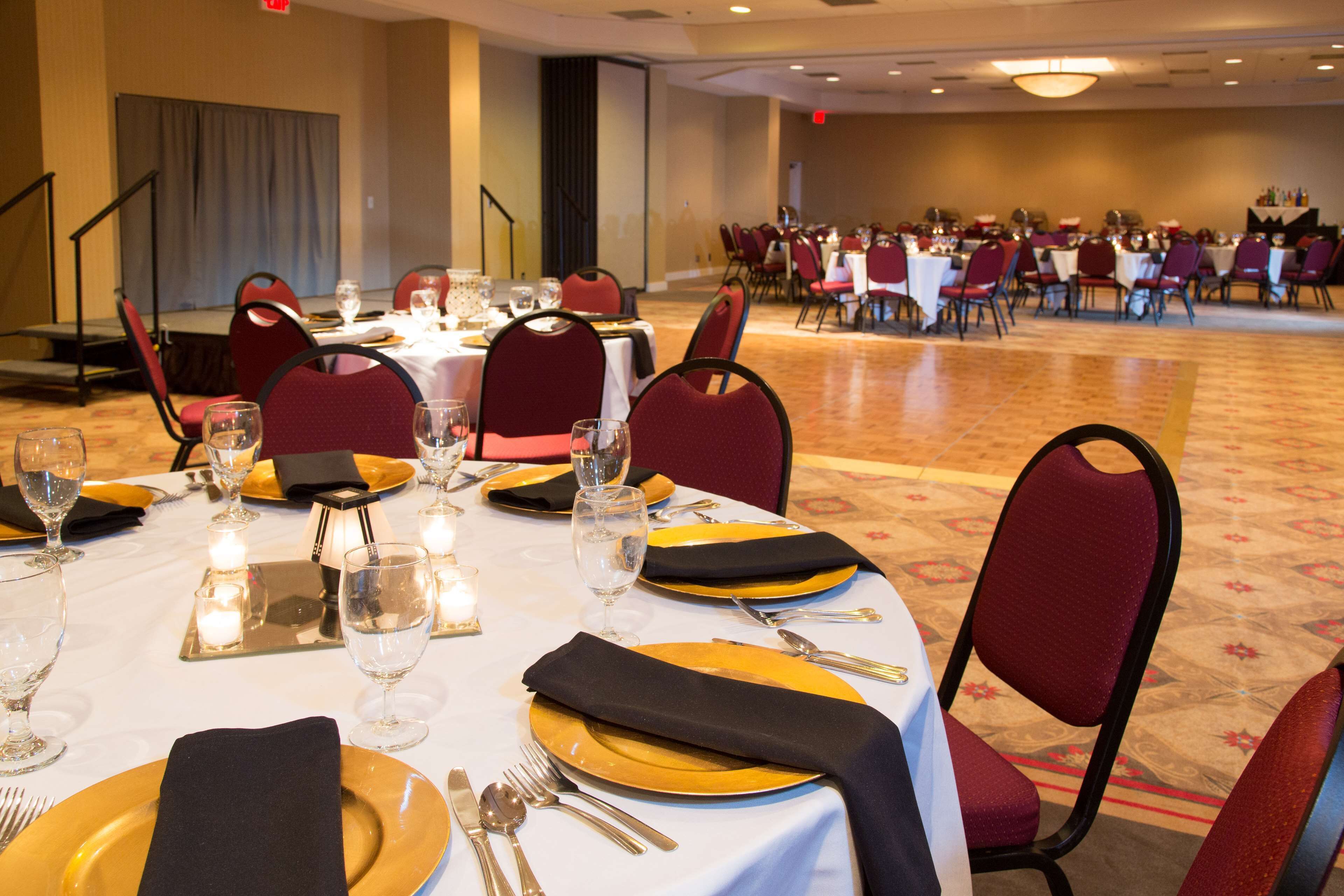 A banquet hall with round tables set for dining, equipped with gold and black tableware, and red upholstered chairs.