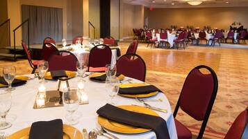 A banquet hall with round tables set for dining, equipped with gold and black tableware, and red upholstered chairs.
