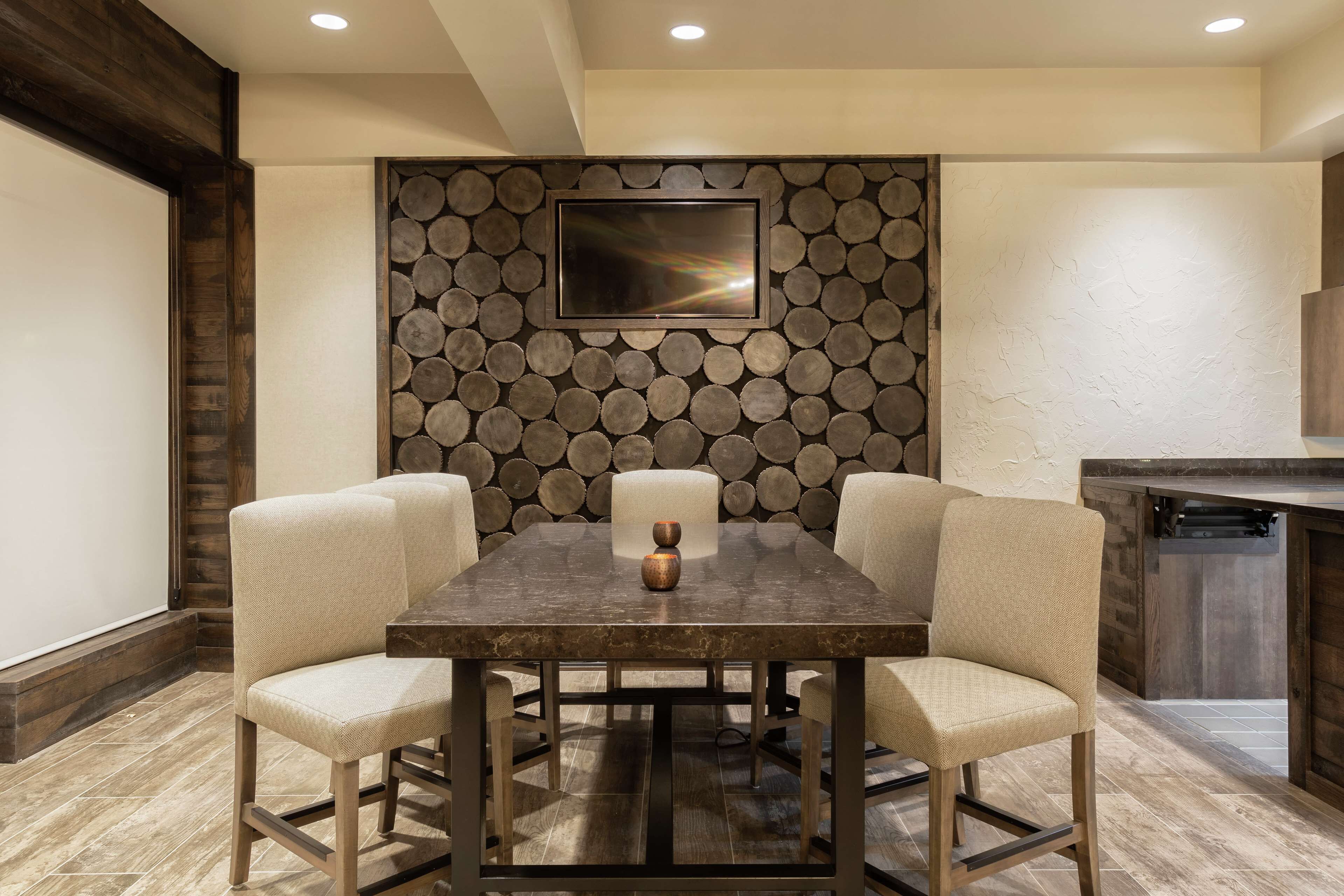 A dining area with a rectangular table, four upholstered chairs, a flat-screen TV mounted on a wall with a circular wood pattern, and a kitchen area with a wooden counter.