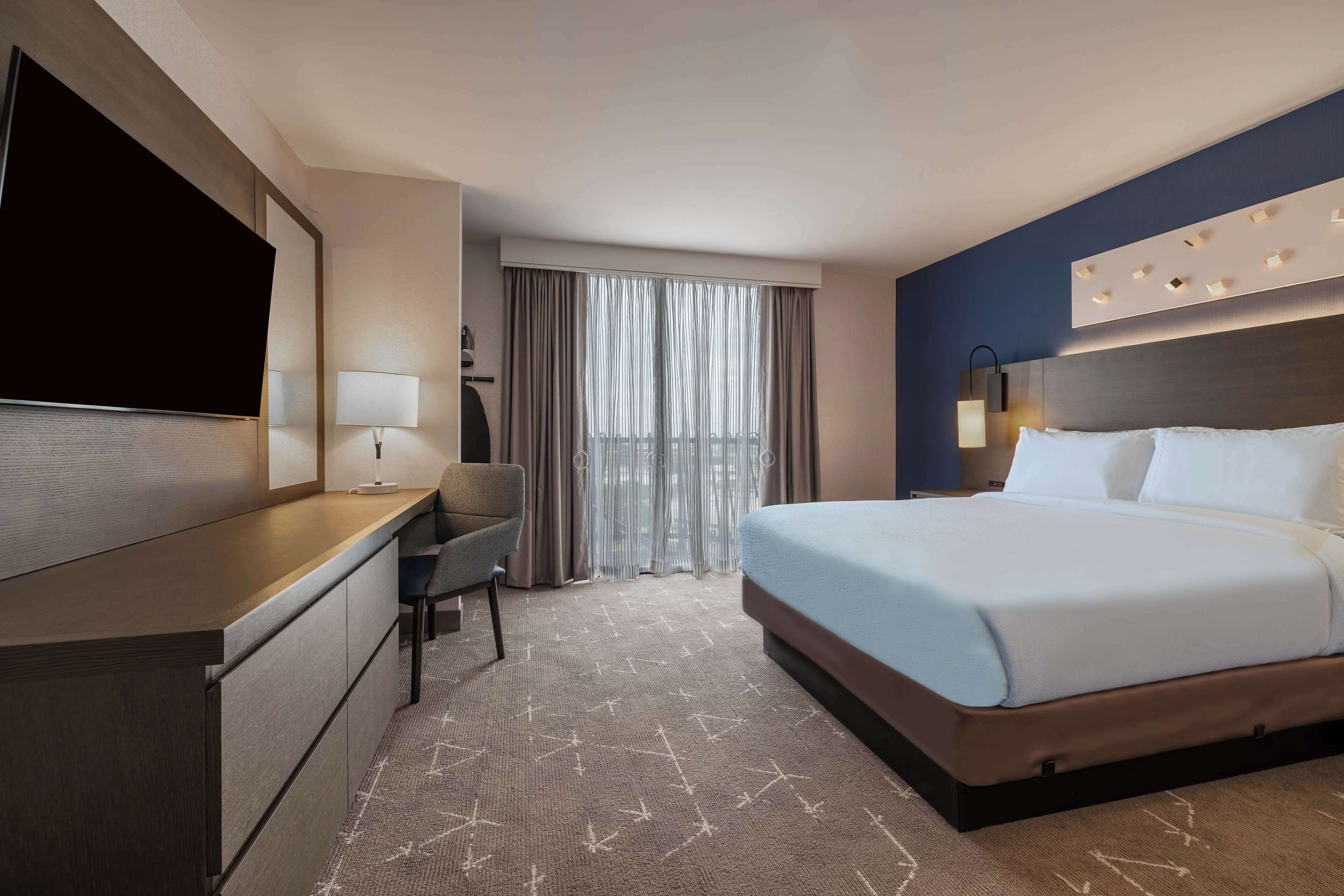 Habitación de hotel con cama, escritorio, silla, televisor y ventana con cortinas.