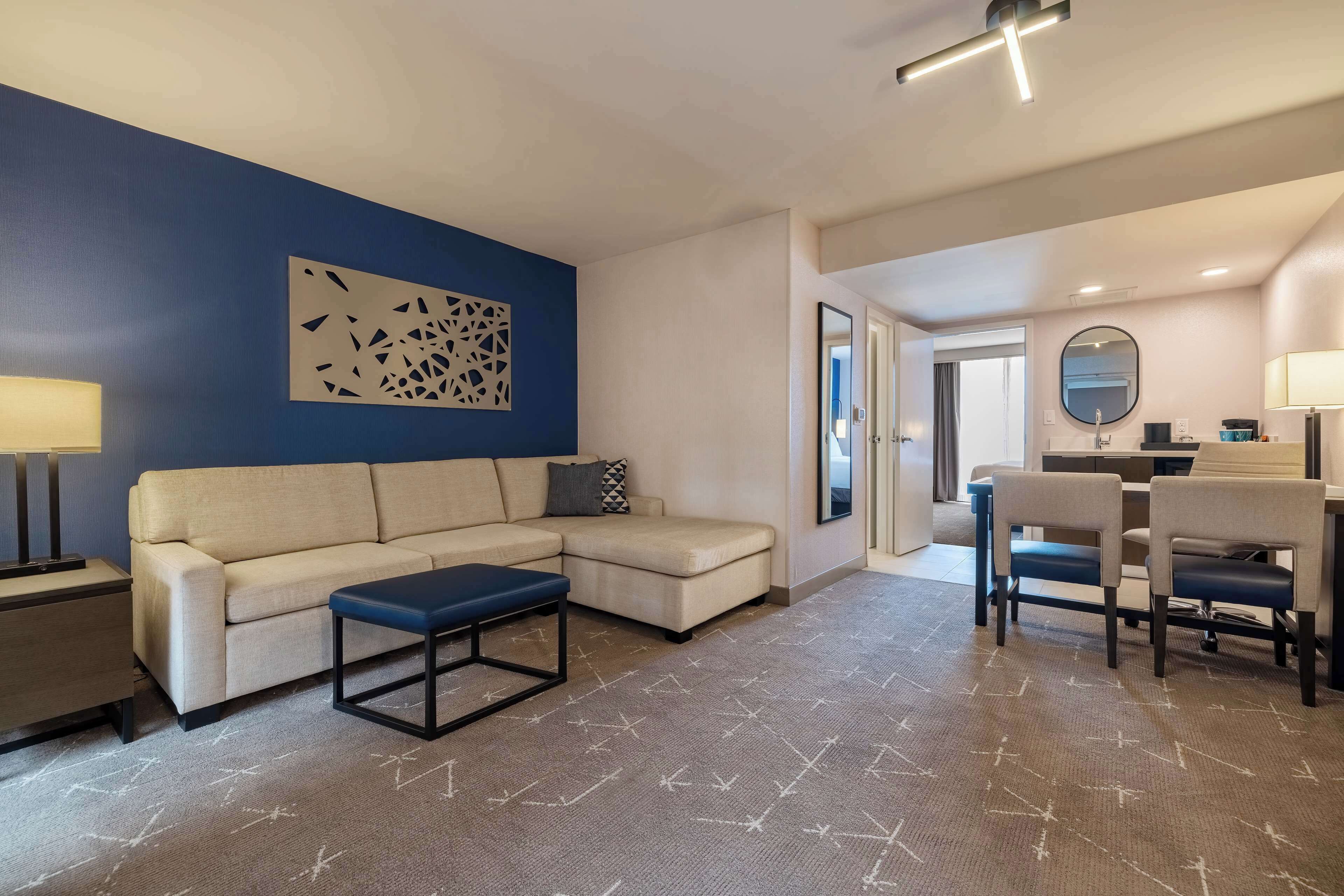 A modern hotel room with a beige sectional sofa, a small blue ottoman, a dining area with two chairs, a table, and a desk with a mirror.